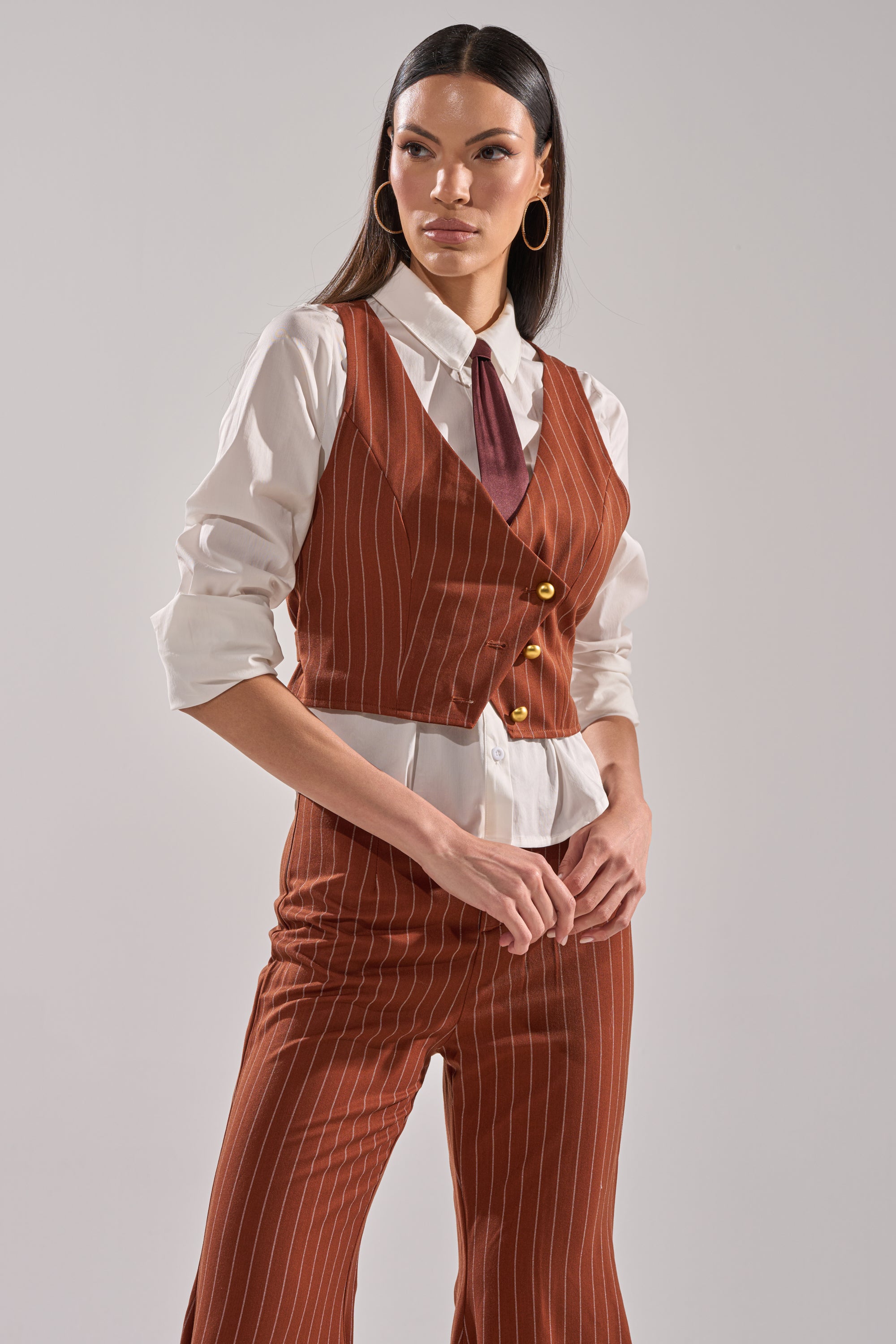 A woman stands confidently in the BUSINESS FIRST VEST, a rust-colored, cropped pinstripe vest with matching pants over a white shirt and burgundy tie against a plain background.