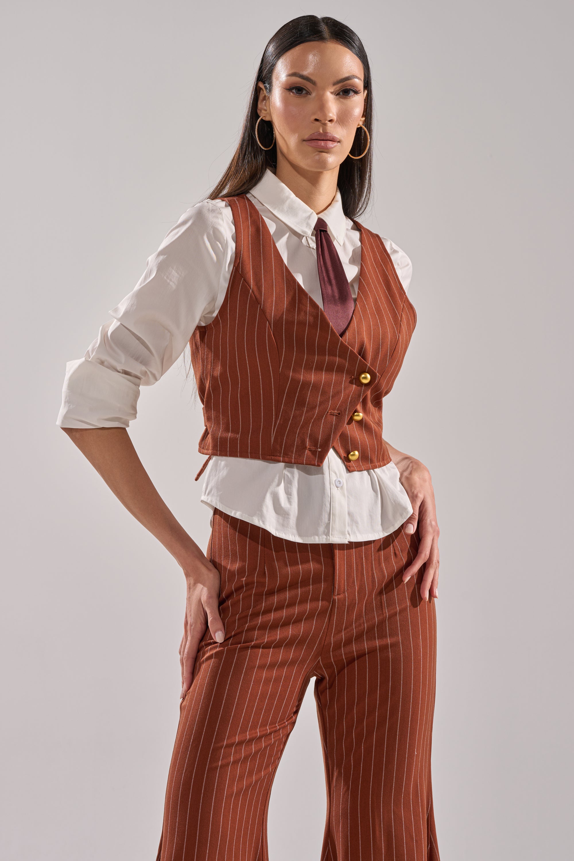A woman poses confidently in the BUSINESS FIRST VEST—a rust pinstripe suit with flared pants—paired over a white shirt with rolled sleeves and a maroon tie, accessorized with large hoop earrings and long, straight hair.