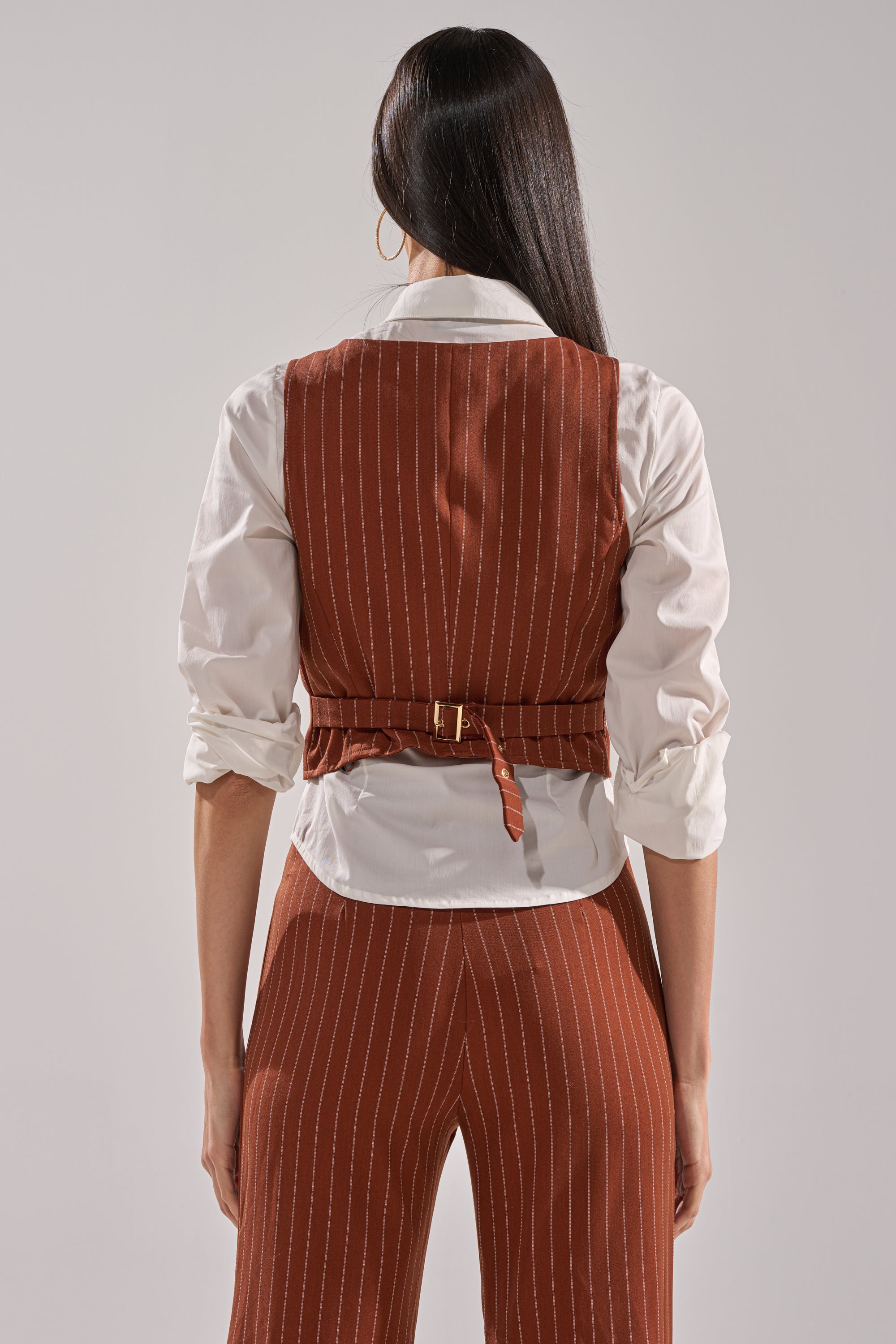 A woman with long dark hair, seen from behind, wears a white shirt and the BUSINESS FIRST VEST—a rust pinstripe vest featuring an adjustable waist belt. Her sleeves are rolled up for a stylish look.