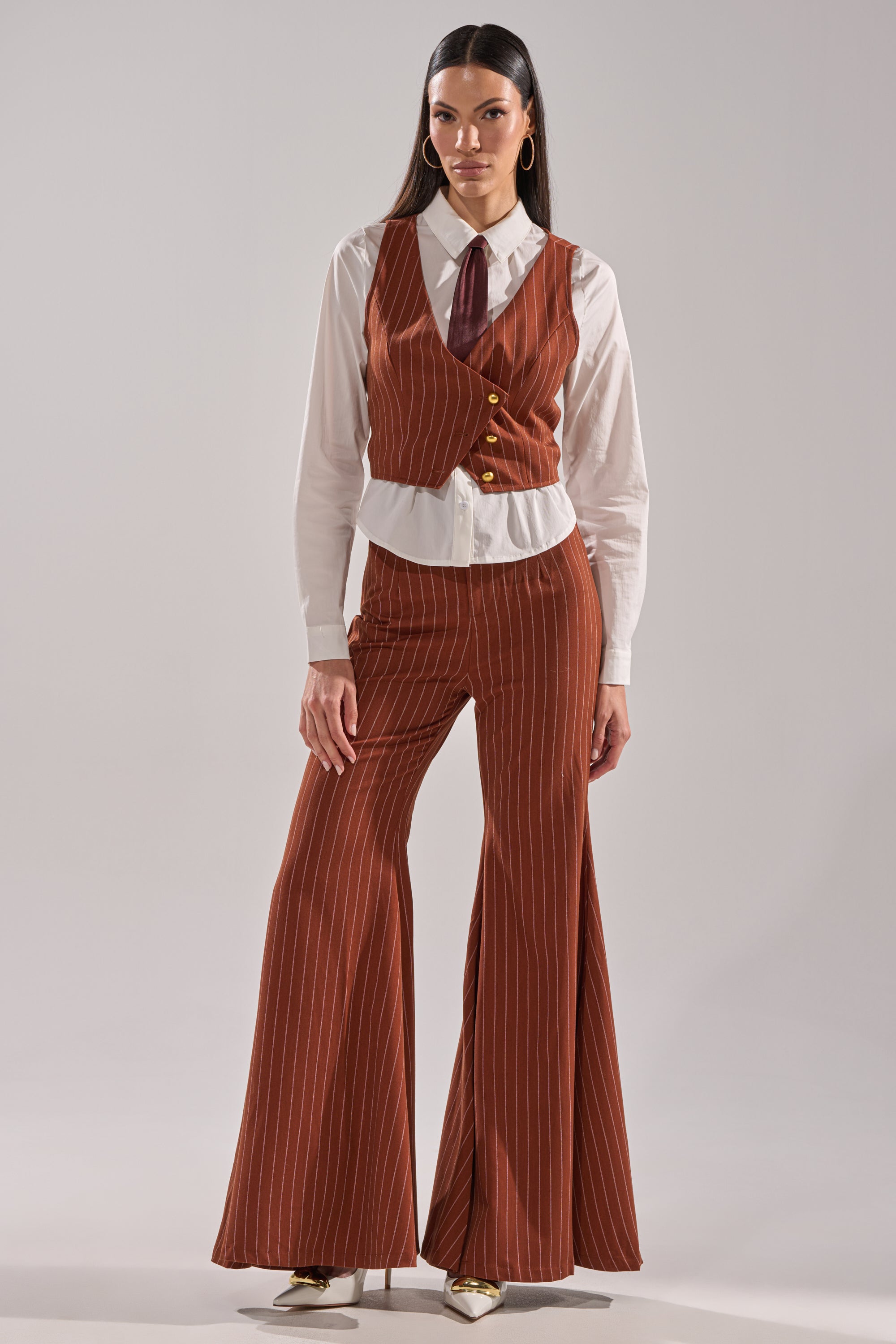 A woman confidently poses against a plain background in the BUSINESS FIRST VEST—brown, pinstriped, and cropped—paired with flared pants, a white button-up shirt, and a maroon necktie.