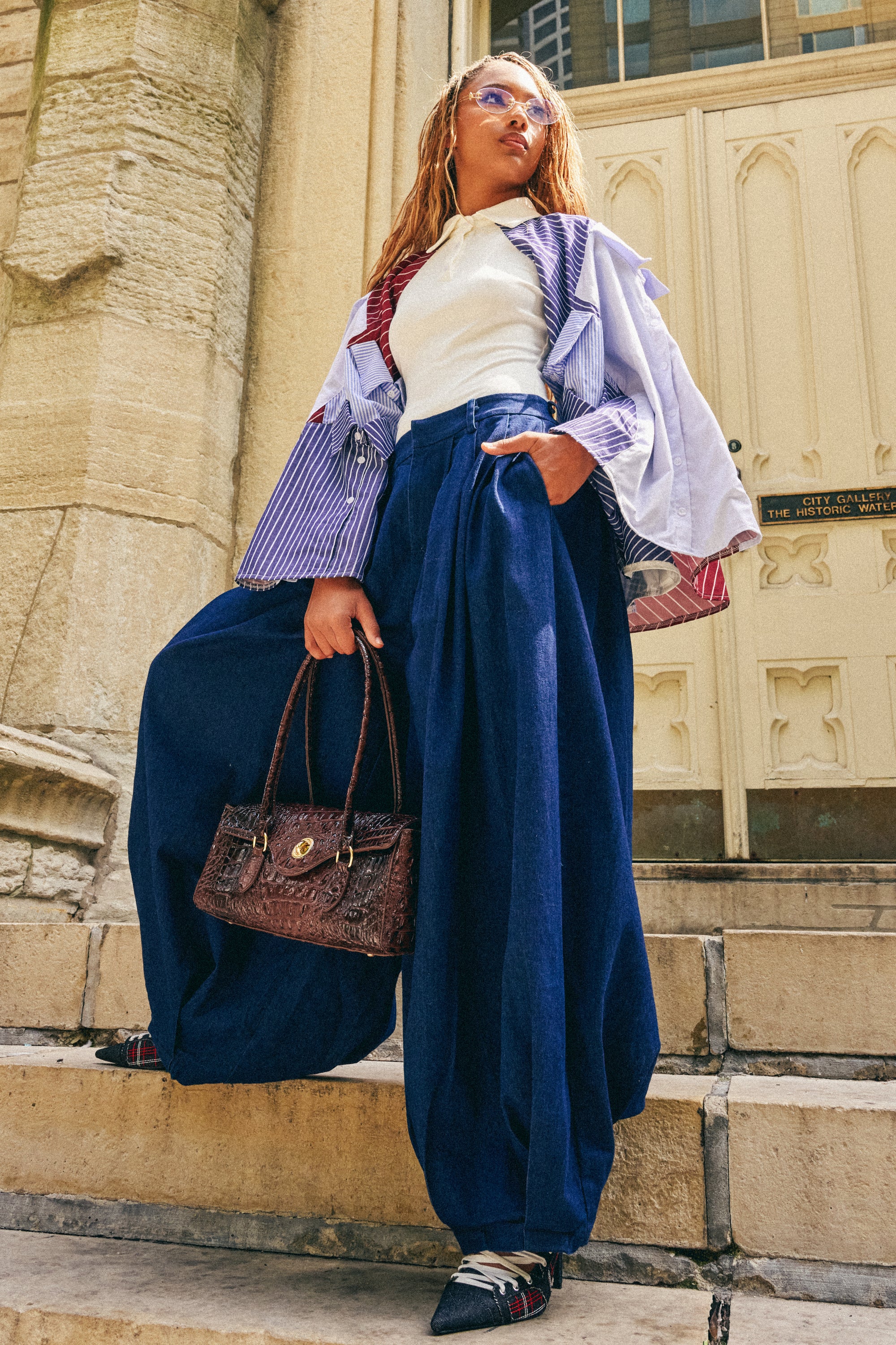 Clad in the BUSINESS IN THE FRONT TROUSER—a chic, wide-leg blue tailored pant—a stylish individual stands on stone steps, complemented by a white top, striped jacket, sunglasses, and a brown handbag. Ornate light-colored doors and elegant stone architecture set the scene in the background.