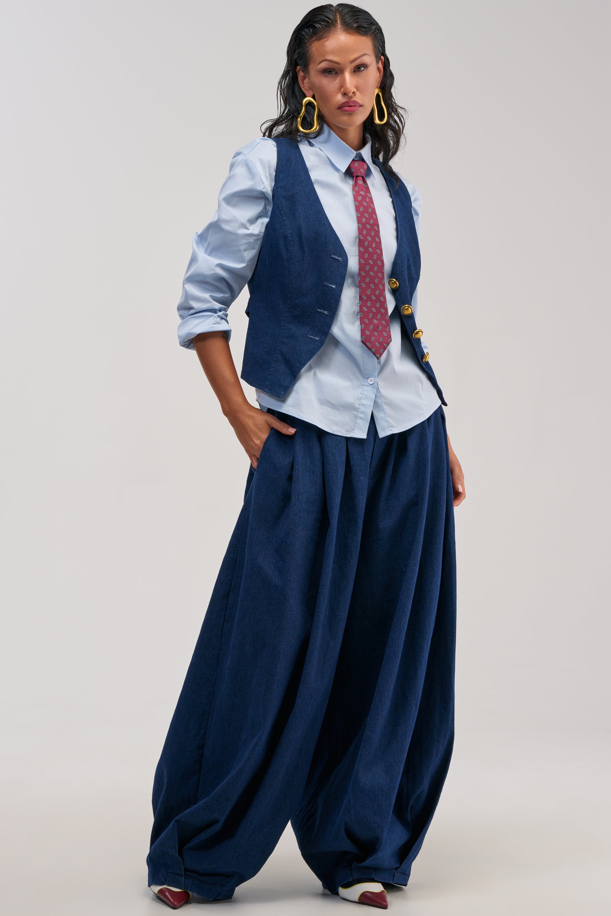 A woman stands confidently in the BUSINESS IN THE FRONT TROUSER—a pair of wide-leg tailored trousers—paired with a blue shirt, red tie, and dark blue vest. She has wavy dark hair, large yellow earrings, and red shoes. The background is plain and light-colored.