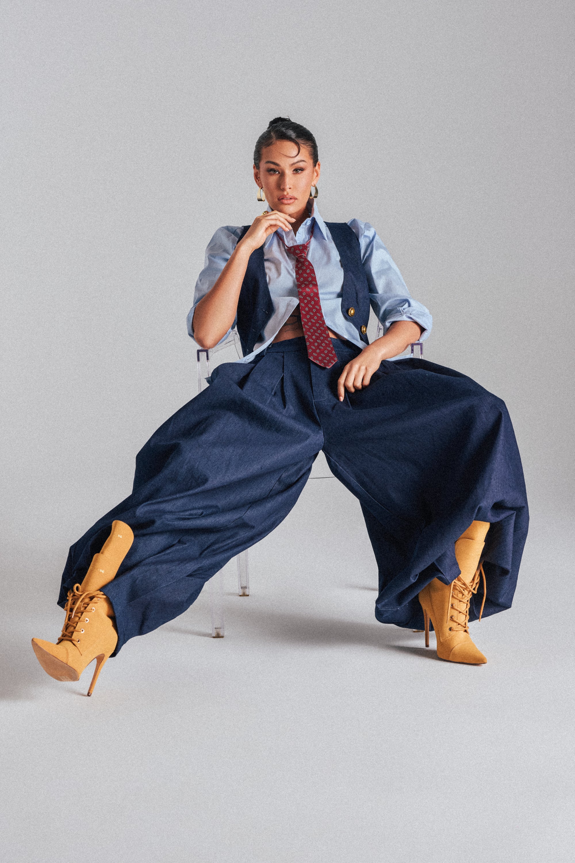 A woman sits confidently on a clear chair against a plain background, wearing BUSINESS IN THE FRONT TROUSER: dark wash, high-rise, wide-leg denim paired with a matching vest, light blue shirt, red patterned tie, and tan high-heeled lace-up boots.