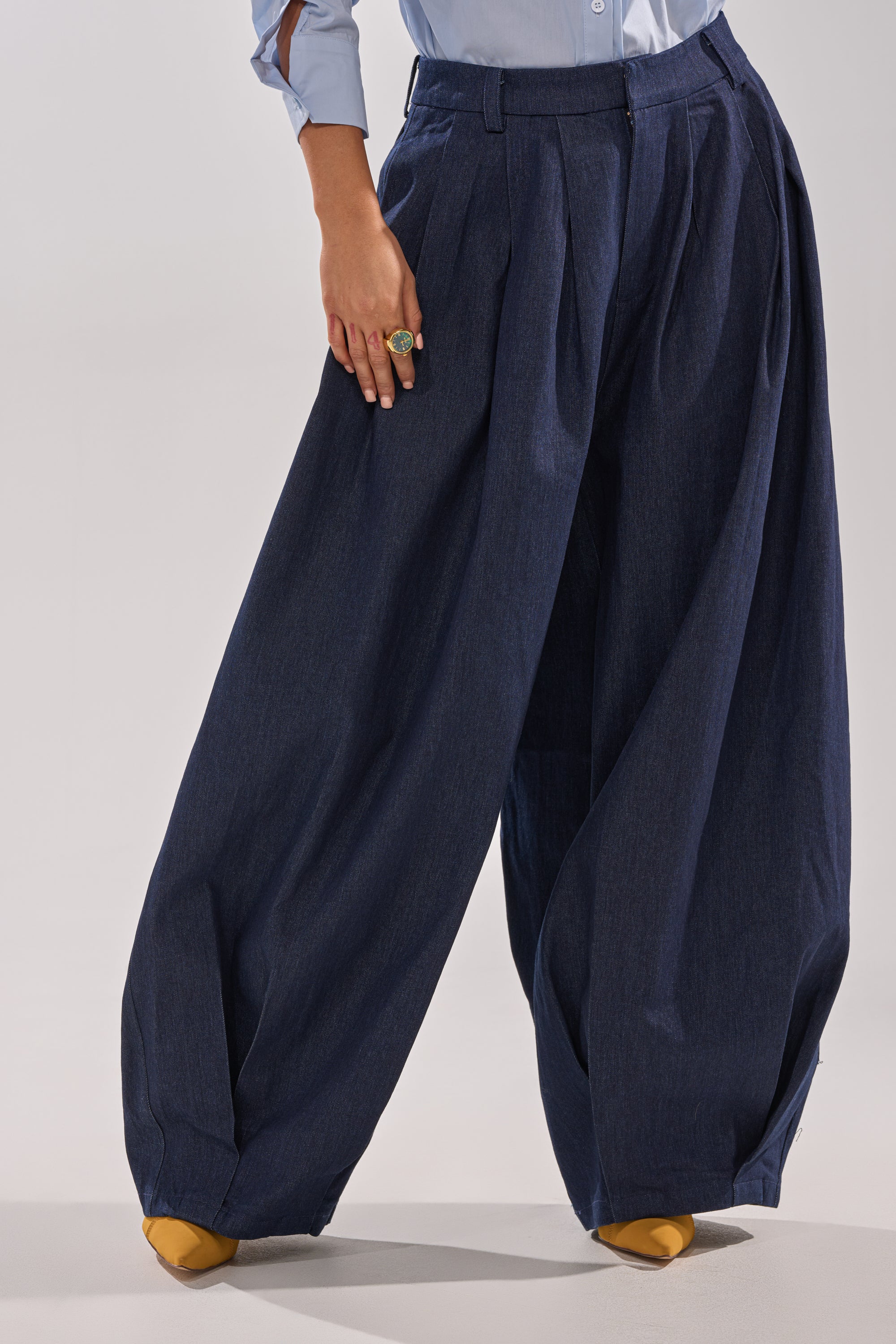 A person is shown from the waist down wearing the BUSINESS IN THE FRONT TROUSER—wide-leg, high-rise dark wash denim with pleats—paired with a light blue shirt and yellow pointed-toe shoes. 4