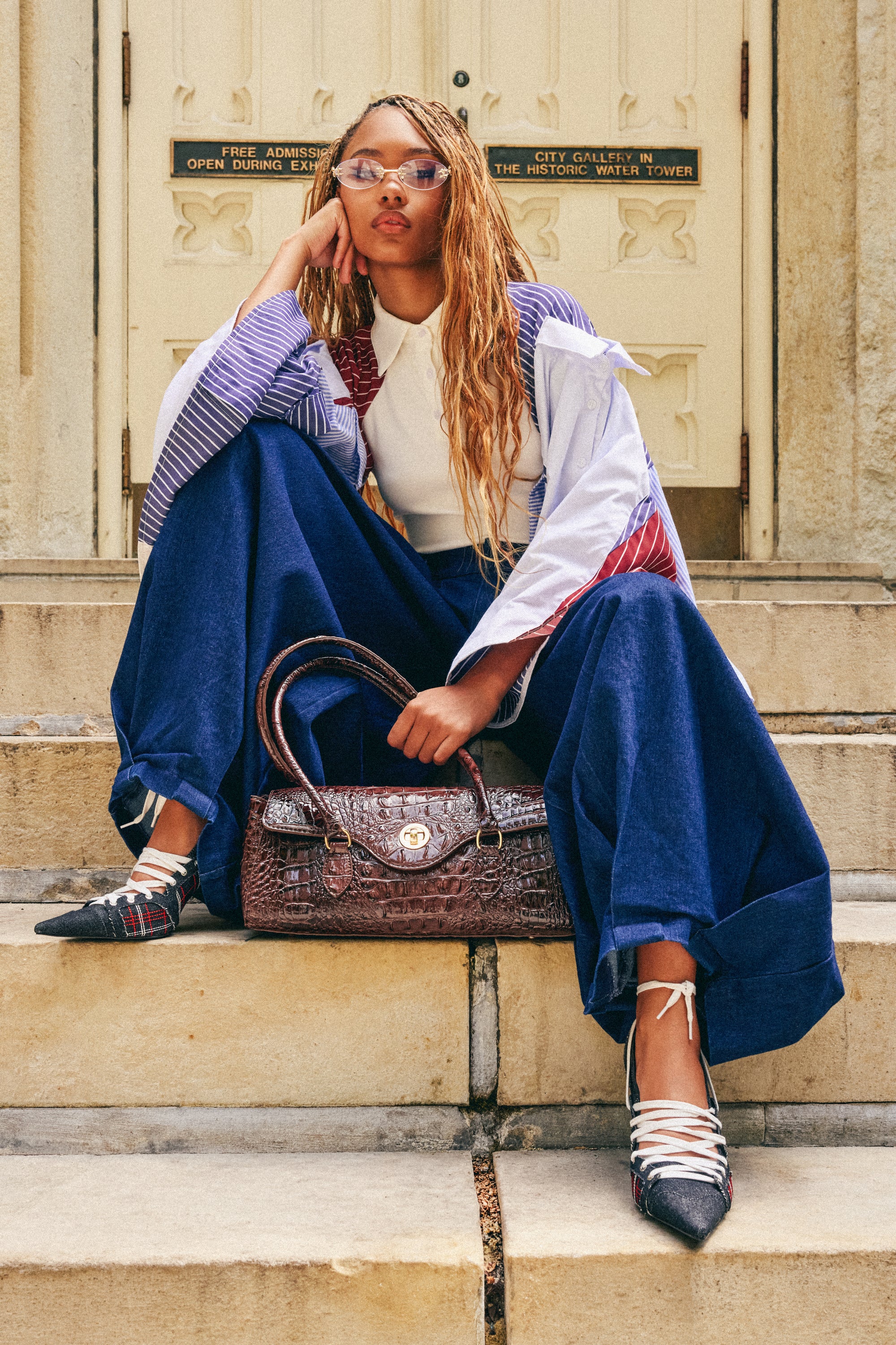 A woman with long braided hair sits on stone steps, wearing the BUSINESS IN THE FRONT TROUSER—oversized, dark-wash, wide-leg denim trousers with a high-rise waist—paired with a white top, layered sleeves, pointed shoes, sunglasses, and holding a textured handbag.
