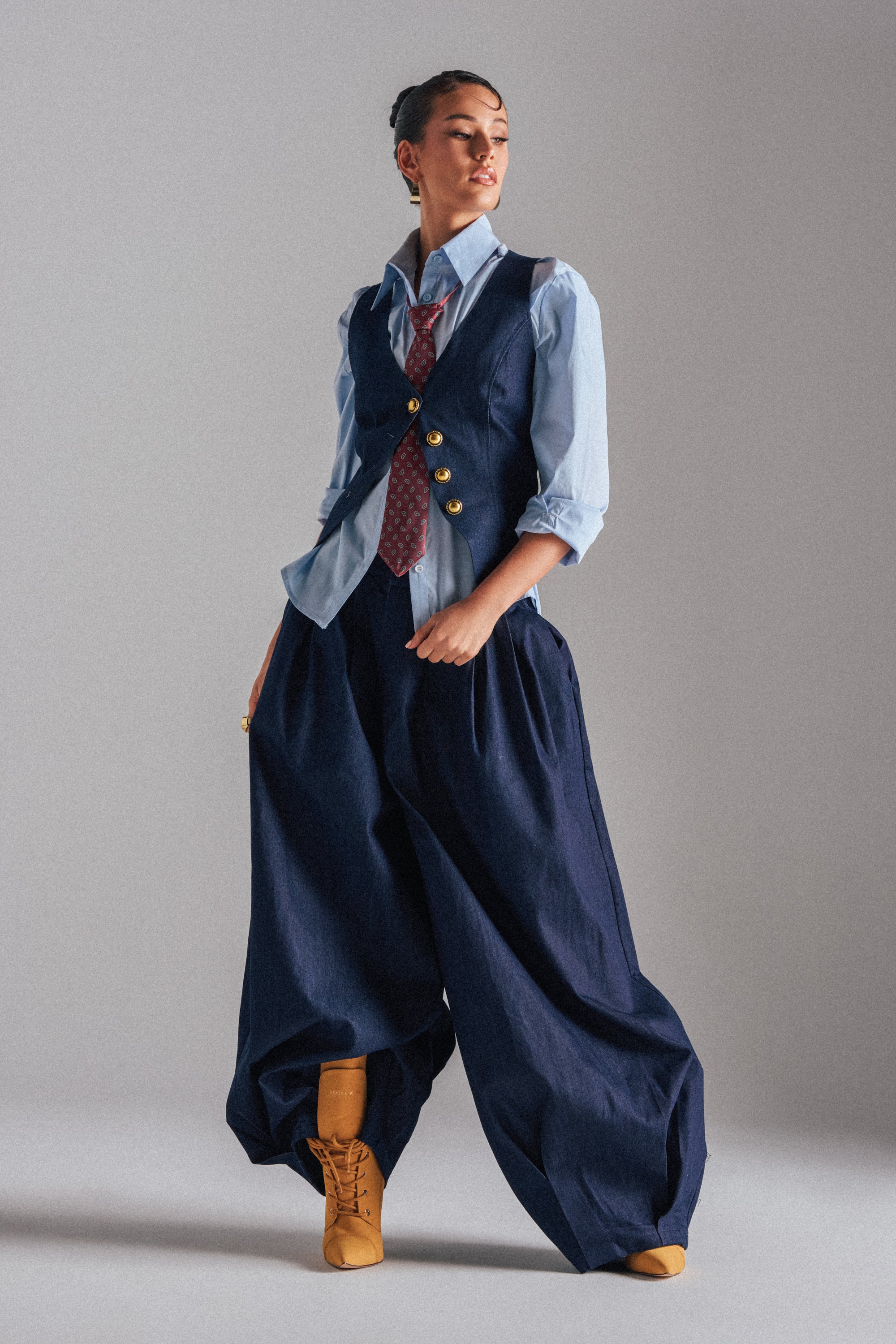 A person poses confidently in the BUSINESS IN THE FRONT TROUSER—wide-leg navy pants with a high-rise waist—paired with a light blue button-up shirt, navy vest with gold buttons, red patterned tie, and tan lace-up boots against a plain gray background. 2
