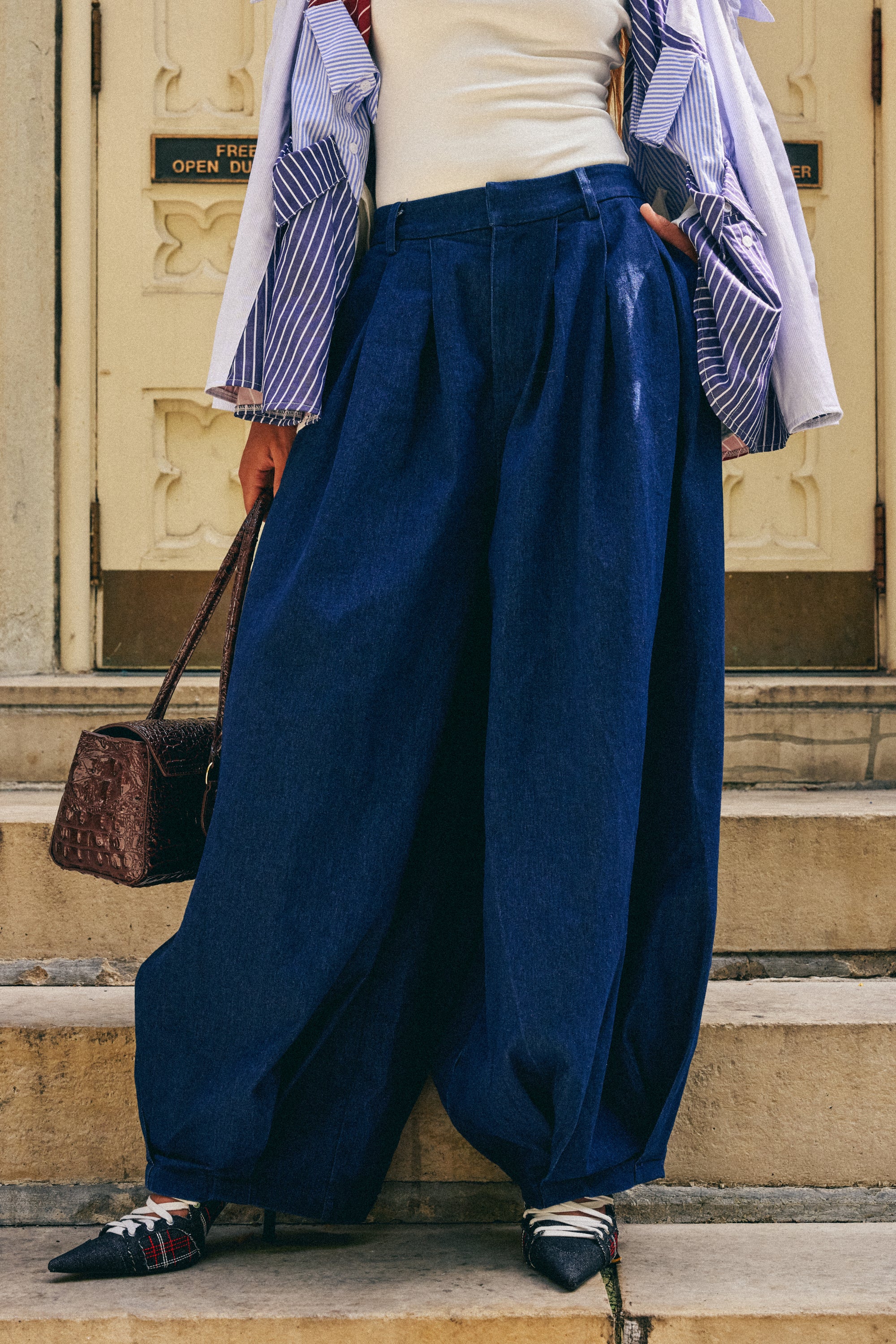 Wearing the BUSINESS IN THE FRONT TROUSER—an oversized, high-rise wide-leg denim—paired with a crisp white top and a striped shirt, this person holds a brown handbag and stands on stone steps before ornate building doors.