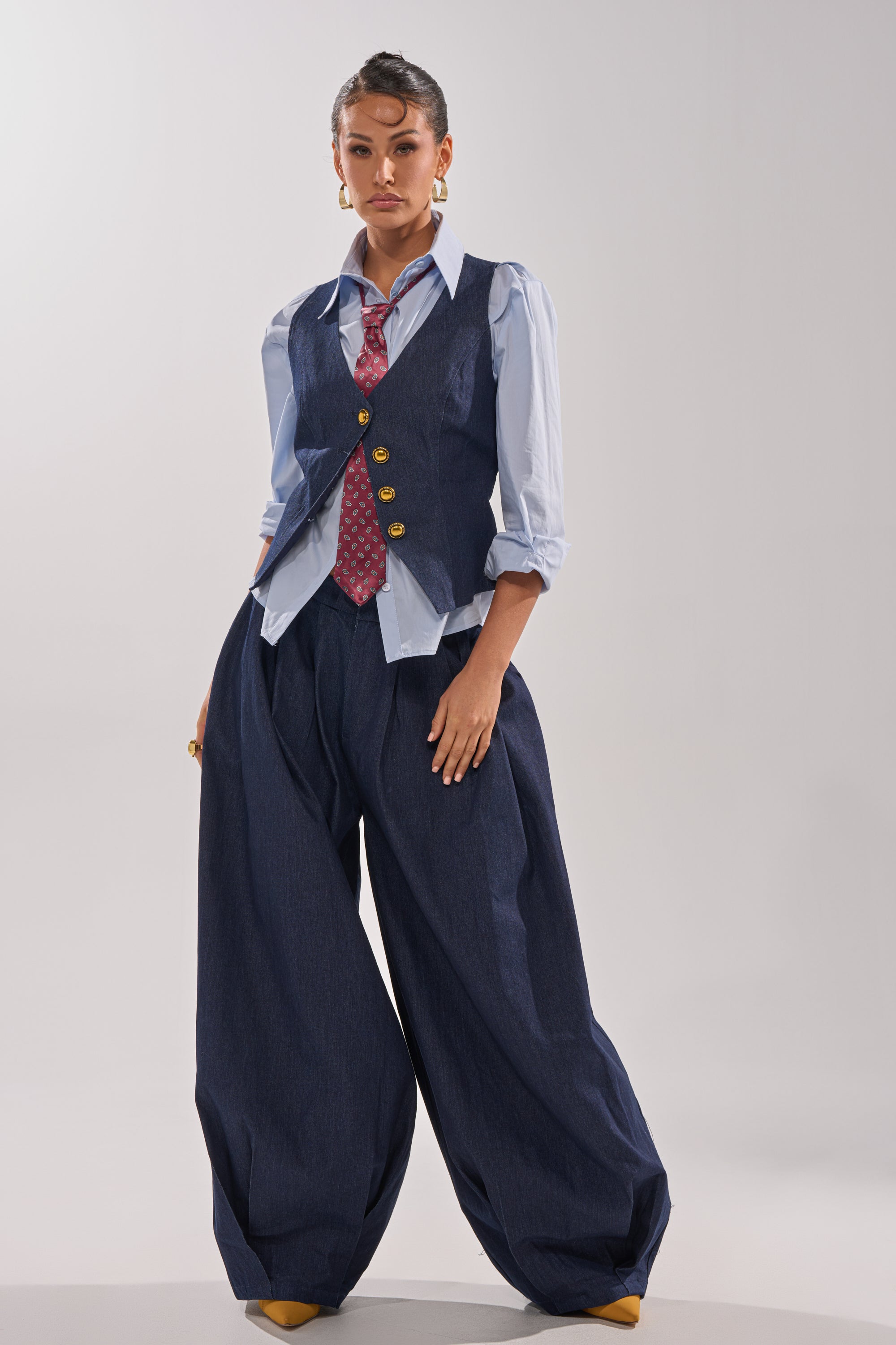A woman stands confidently in BUSINESS IN THE FRONT TROUSER, dark wash wide-leg denim with a high-rise waist, paired with a light blue shirt, blue vest with gold buttons, red patterned tie, gold earrings, and yellow shoes against a plain light background. 8