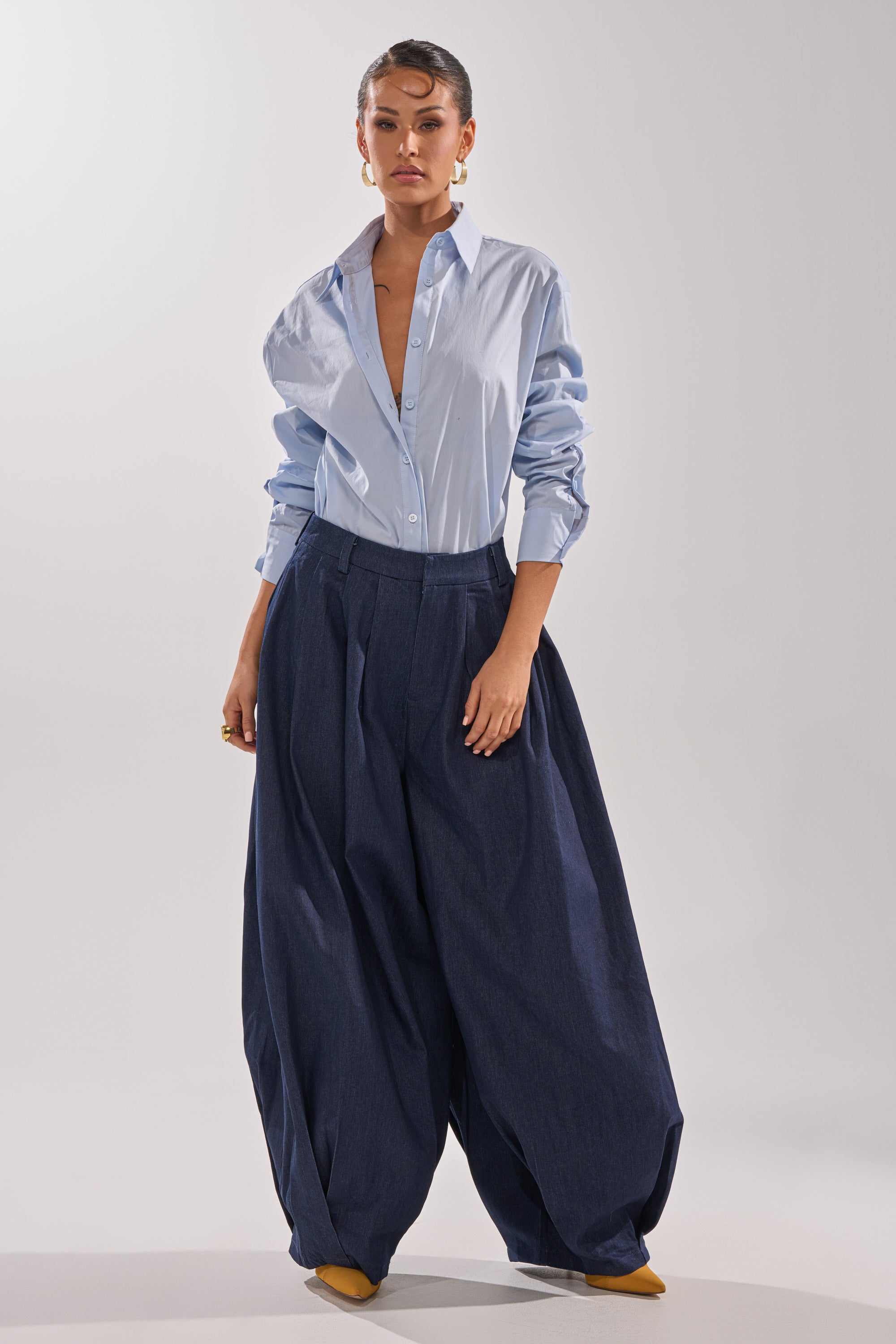 A woman poses against a plain light background wearing navy blue BUSINESS IN THE FRONT TROUSER—high-rise, wide-leg style—with a light blue button-up shirt, rolled sleeves, gold earrings, and yellow pointed-toe shoes. 10
