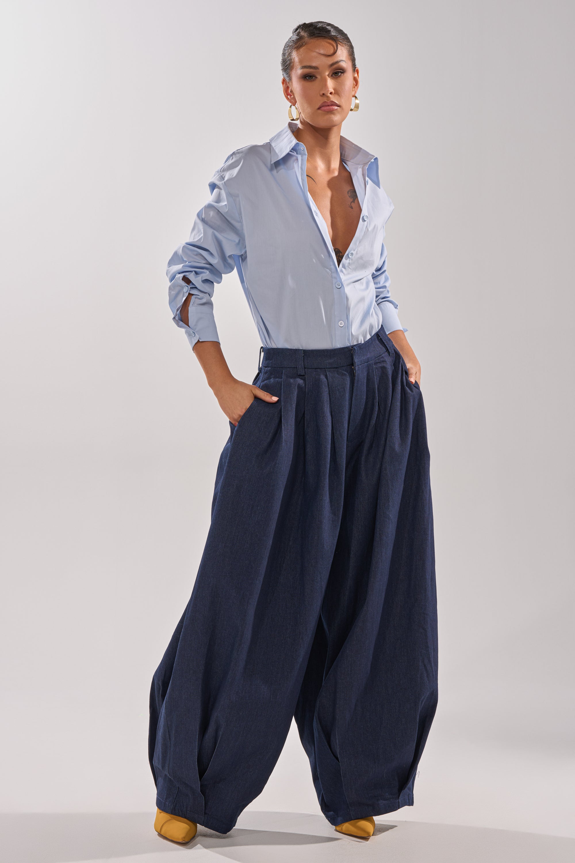 A person poses confidently in wide-leg BUSINESS IN THE FRONT TROUSER, paired with a light blue button-down shirt and mustard yellow shoes, standing against a plain light background. 11