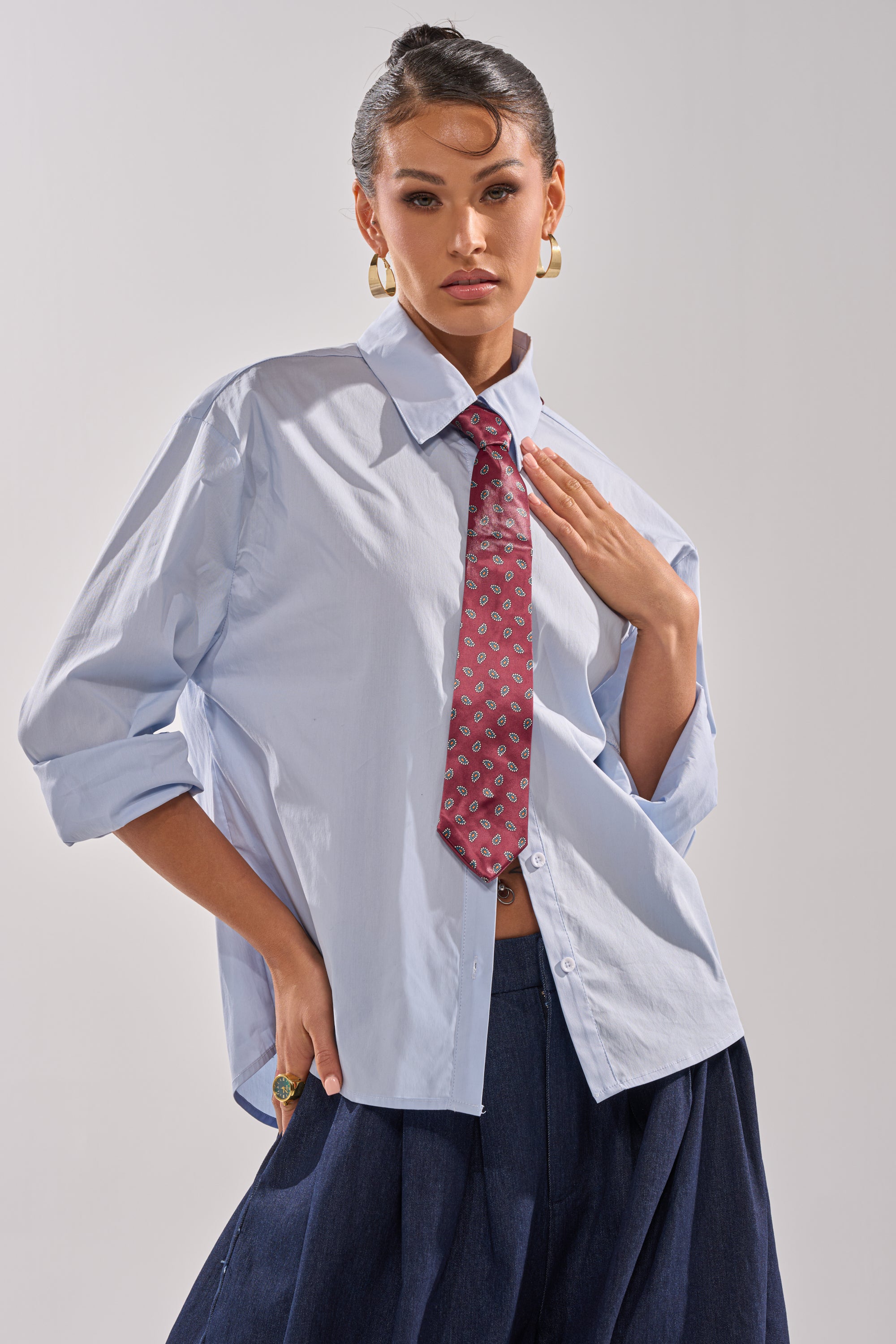 A person poses confidently against a plain background wearing the BUSINESS IN THE FRONT BUTTON DOWN, a loose blue blouse with a tailored collar, paired with a patterned red tie, wide-leg dark pants, gold hoop earrings, and a ring.