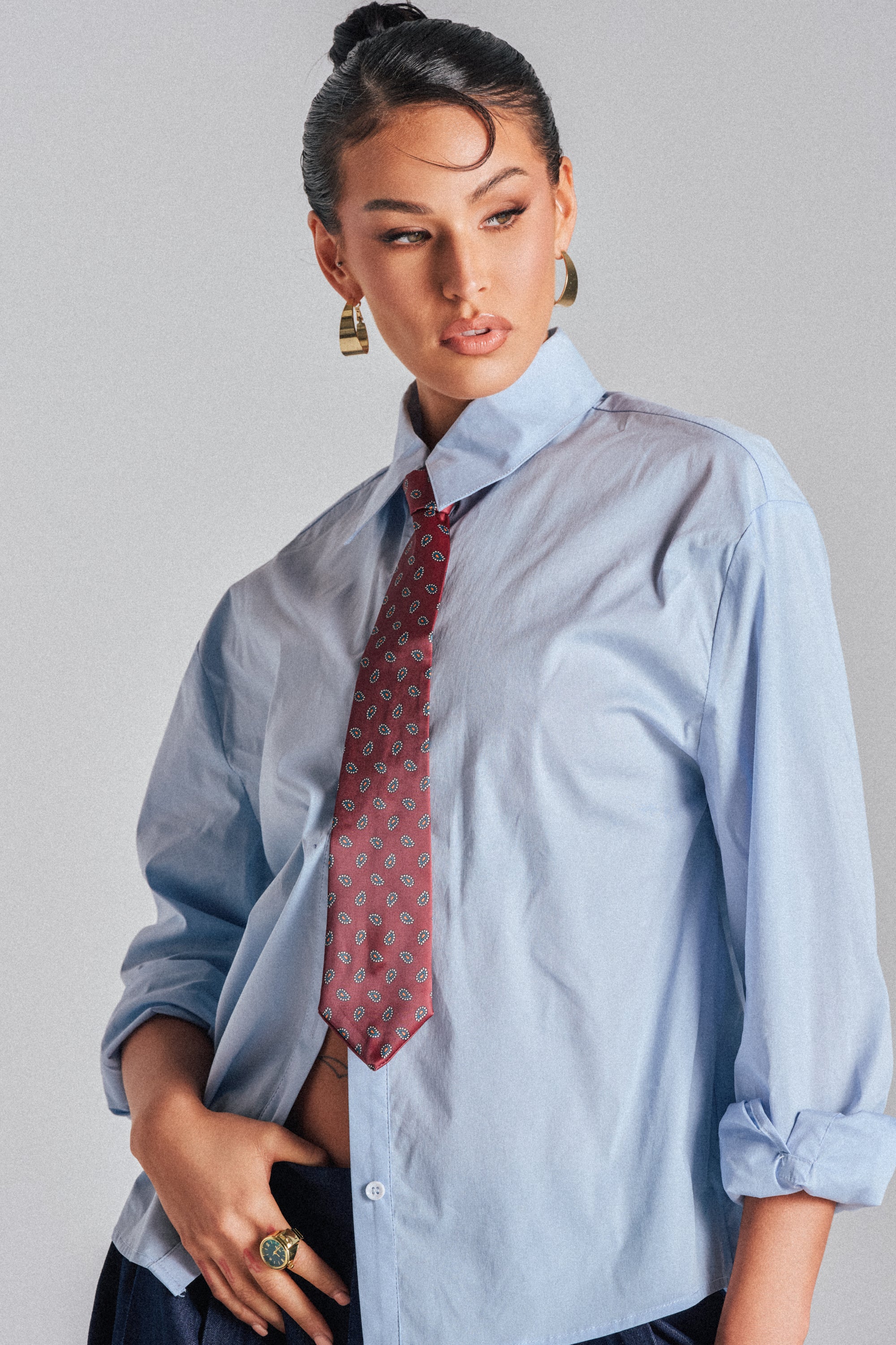 A woman with dark hair in a bun wears the BUSINESS IN THE FRONT BUTTON DOWN, paired with a red tie, gold hoop earrings, and a green ring. She poses confidently, looking to the side with one hand on her hip—perfect for the office.