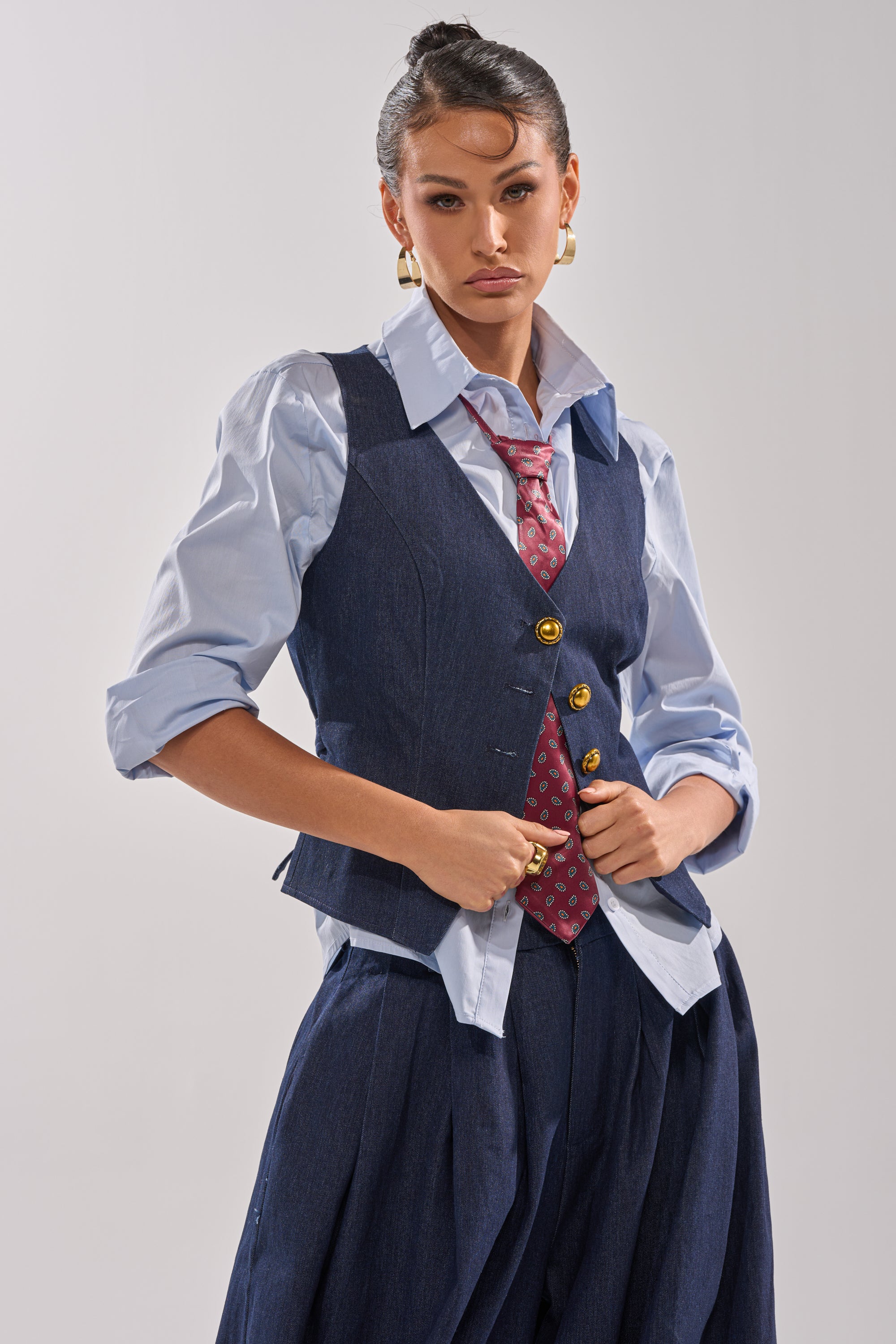 A person with slicked-back hair wears the BUSINESS IN THE FRONT BUTTON DOWN—a blue tailored-collar blouse with rolled sleeves—paired with a dark blue vest, red tie, pleated pants, and gold hoop earrings, posing confidently.