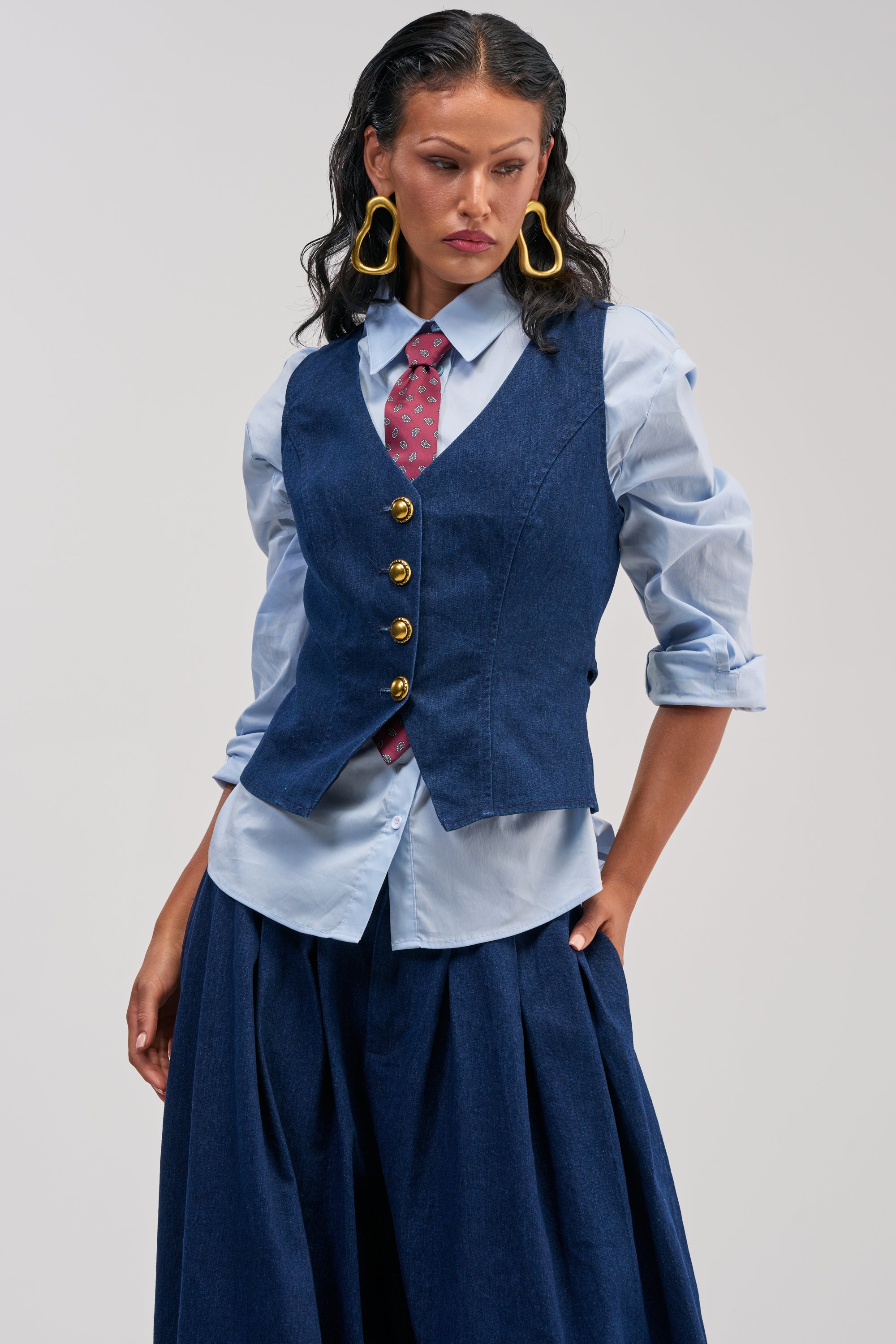 Wearing the “BUSINESS IN THE FRONT BUTTON DOWN,” a woman layers a blue buttoned vest over a light blue shirt styled with a removable red patterned tie, pairing it with wide-leg navy trousers and bold yellow earrings for a smart-casual look as she poses against a plain background.