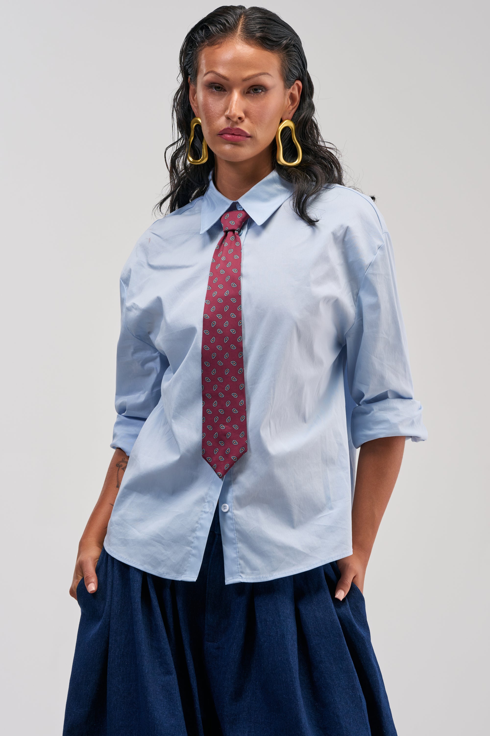 A woman with long dark hair models the BUSINESS IN THE FRONT BUTTON DOWN in light blue, paired with a red patterned removable tie, a dark blue skirt, and bold yellow statement earrings. Her rolled-up sleeves and confident stance against a plain backdrop highlight this shirt’s versatility for smart-casual fashion.