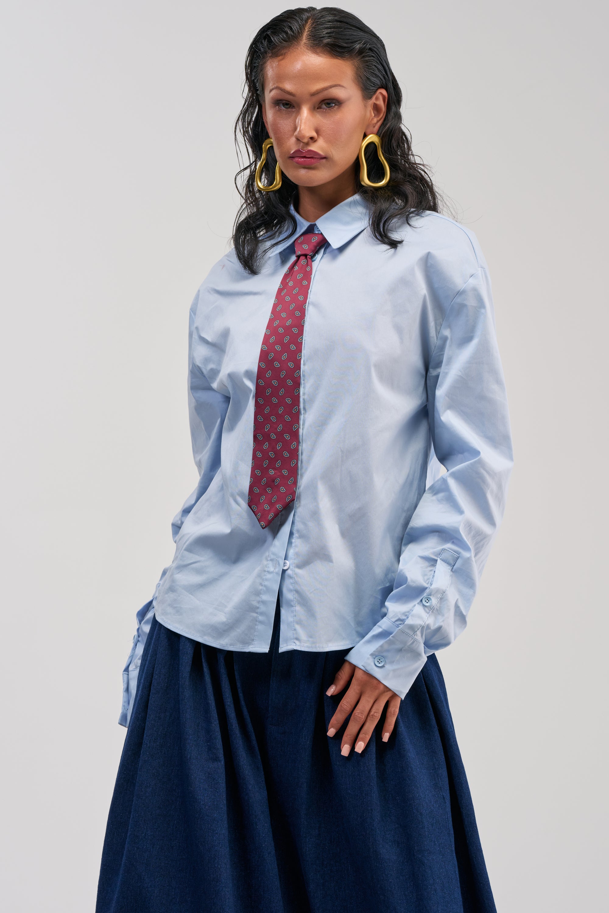 Wearing the BUSINESS IN THE FRONT BUTTON DOWN in light blue, paired with a red patterned removable tie, dark blue skirt, and large yellow earrings, a person with long dark hair stands against a plain background—a perfect example of smart-casual fashion.