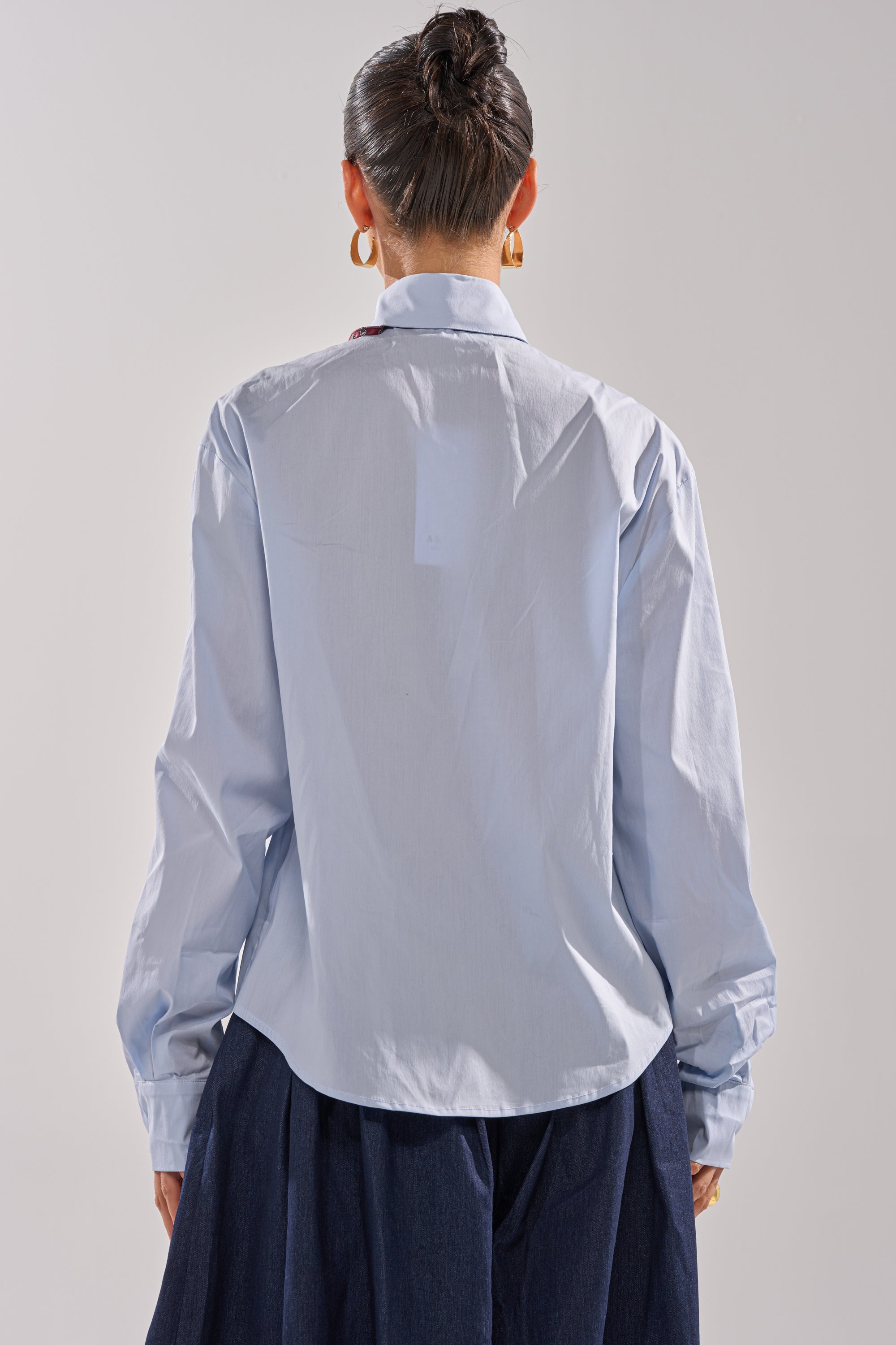 Wearing the BUSINESS IN THE FRONT BUTTON DOWN, a woman with dark hair in a bun and gold hoop earrings stands with her back to the camera against a light background—offering timeless office wear inspiration.