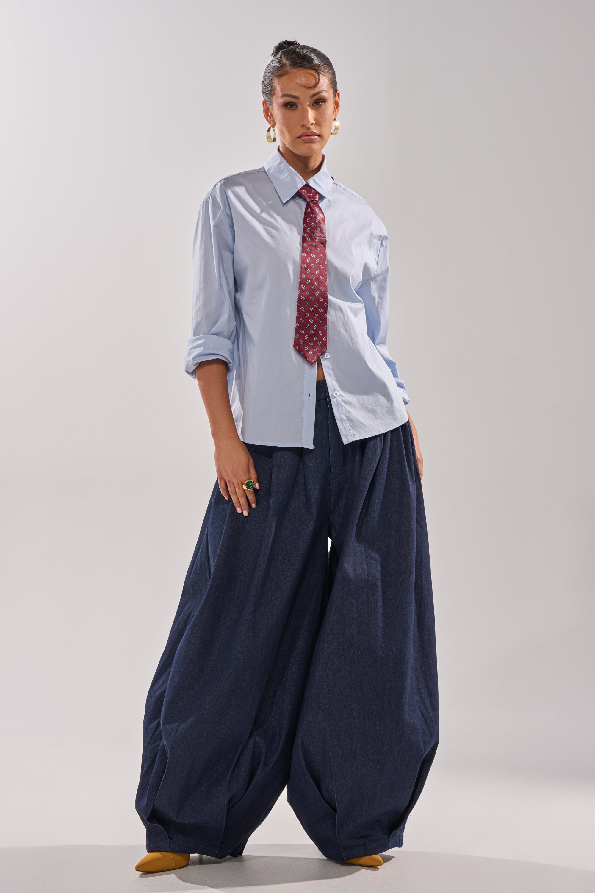 Wearing the BUSINESS IN THE FRONT BUTTON DOWN, a light blue blouse with a tailored collar, paired with red tie, oversized navy wide-leg pants, and yellow shoes—a standout look for bold office style against a simple backdrop.