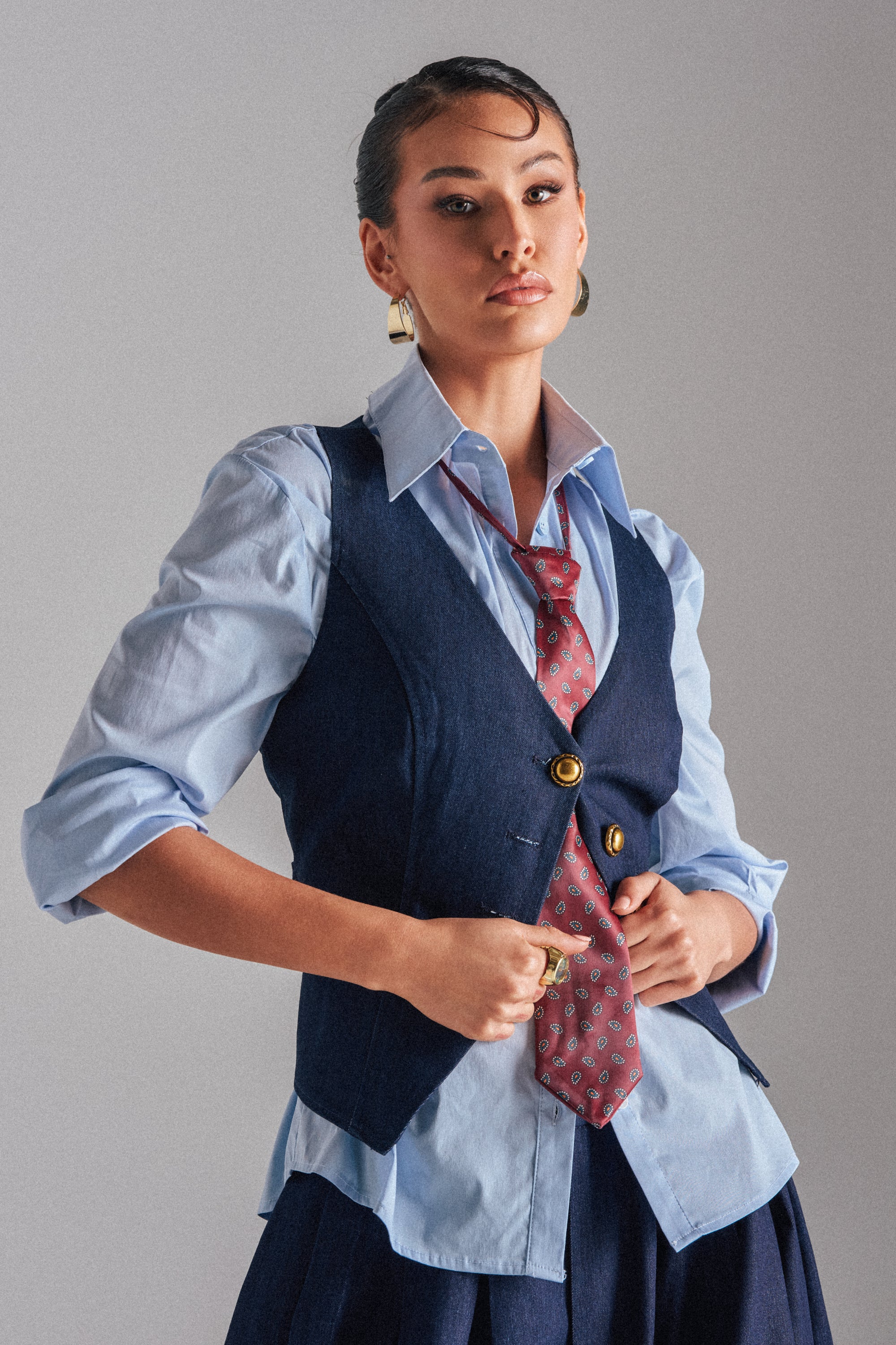 Wearing the BUSINESS IN THE FRONT VEST over a blue button-up with rolled sleeves, a patterned red tie, and hoop earrings, this person stands confidently against a plain background, looking directly at the camera.