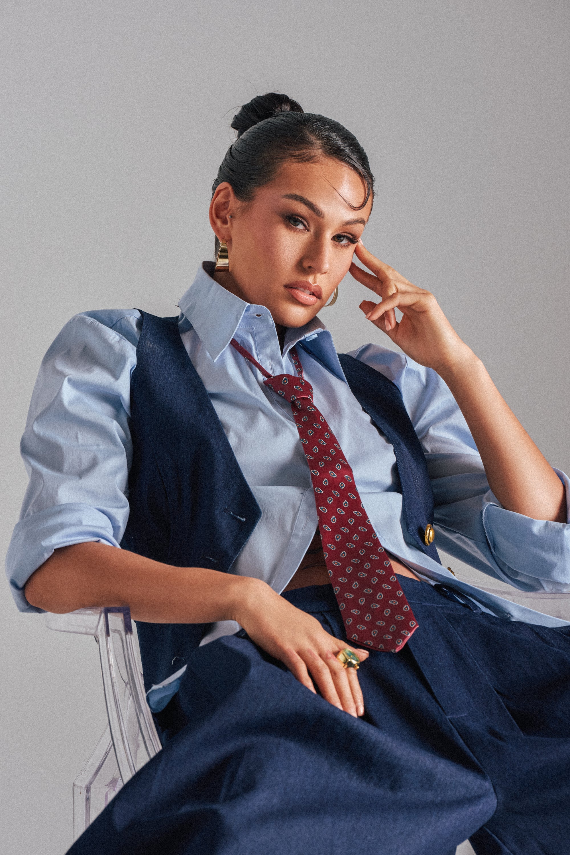 A woman with dark hair in a bun sits on a clear chair wearing the BUSINESS IN THE FRONT VEST over a blue shirt, complemented by a patterned red tie and navy trousers. She rests her head on one hand, looking confidently at the camera.