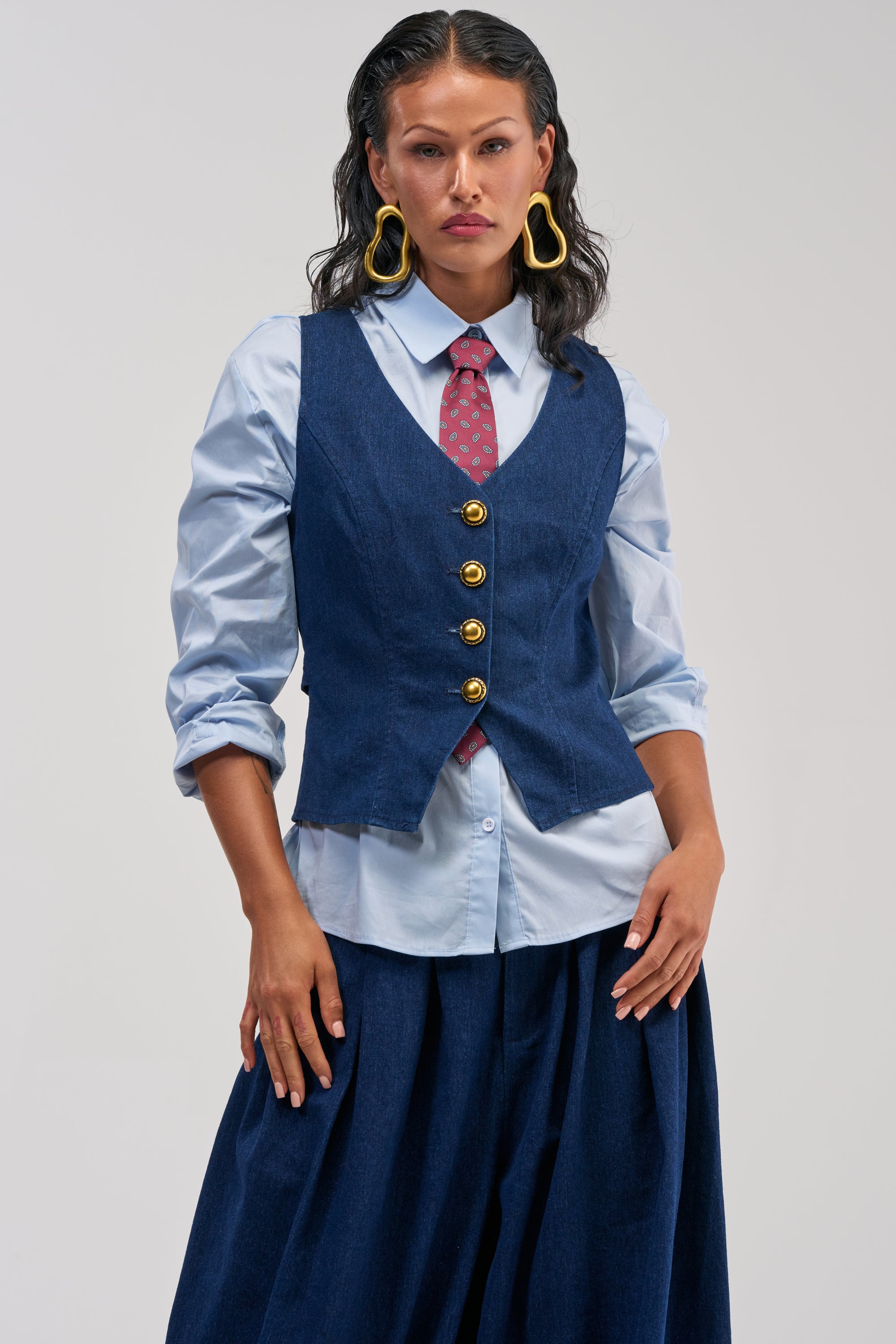 Wearing the BUSINESS IN THE FRONT VEST—a tailored denim piece adorned with gold buttons—a woman with long dark hair pairs it with a light blue button-up shirt, a patterned red tie, a dark blue skirt, and bold yellow earrings. Facing forward with a neutral expression, her ensemble exemplifies smart-casual styling with an understated edge.
