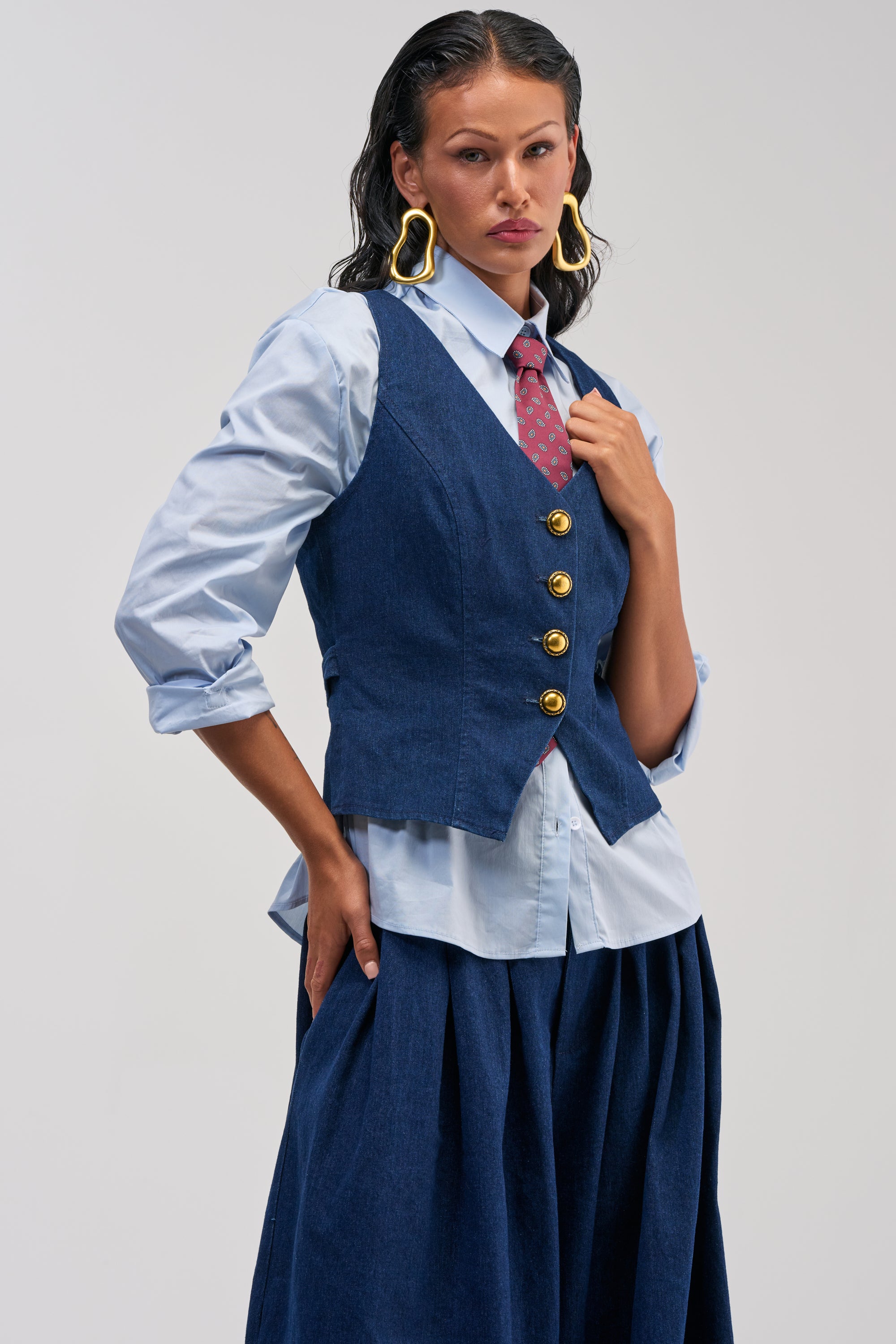 A person poses confidently against a plain background, effortlessly showcasing smart-casual style in the BUSINESS IN THE FRONT VEST—a tailored denim piece with gold buttons—paired with a light blue shirt, red polka-dot tie, blue skirt, and large gold earrings.