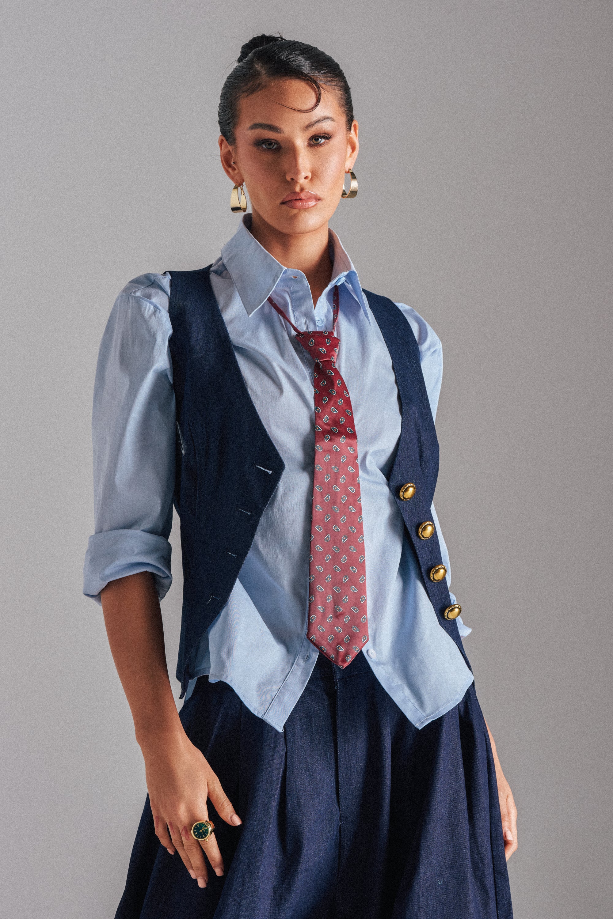 Wearing the BUSINESS IN THE FRONT VEST with gold buttons, a light blue button-up shirt, patterned red tie, dark skirt, gold hoop earrings, and a green ring, this person stands against a neutral background.
