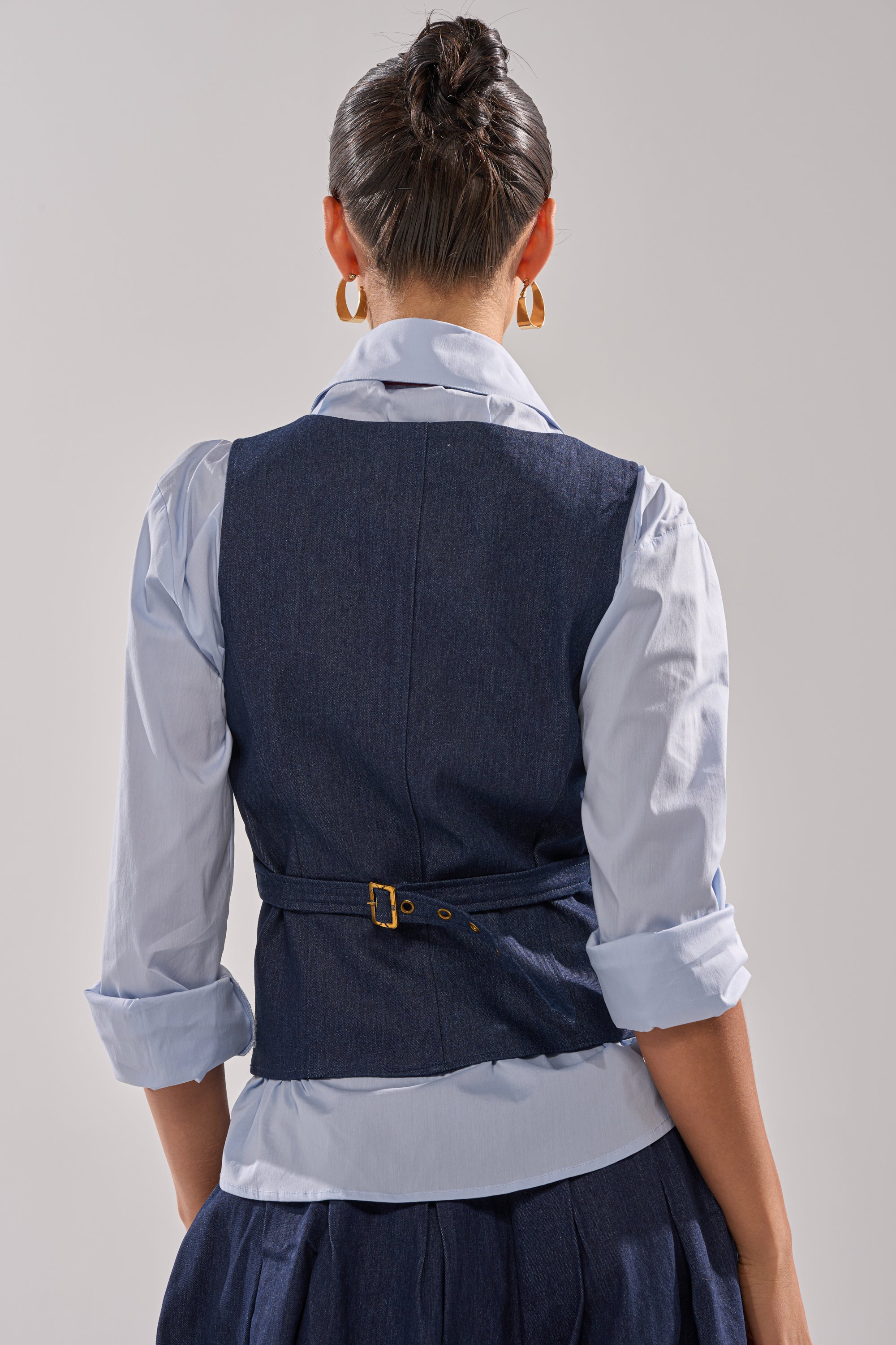 A woman faces away, wearing the BUSINESS IN THE FRONT VEST—a blue denim vest with a tailored fit—over a light blue collared shirt and matching skirt, her hair tied up and accessorized with gold hoop earrings.