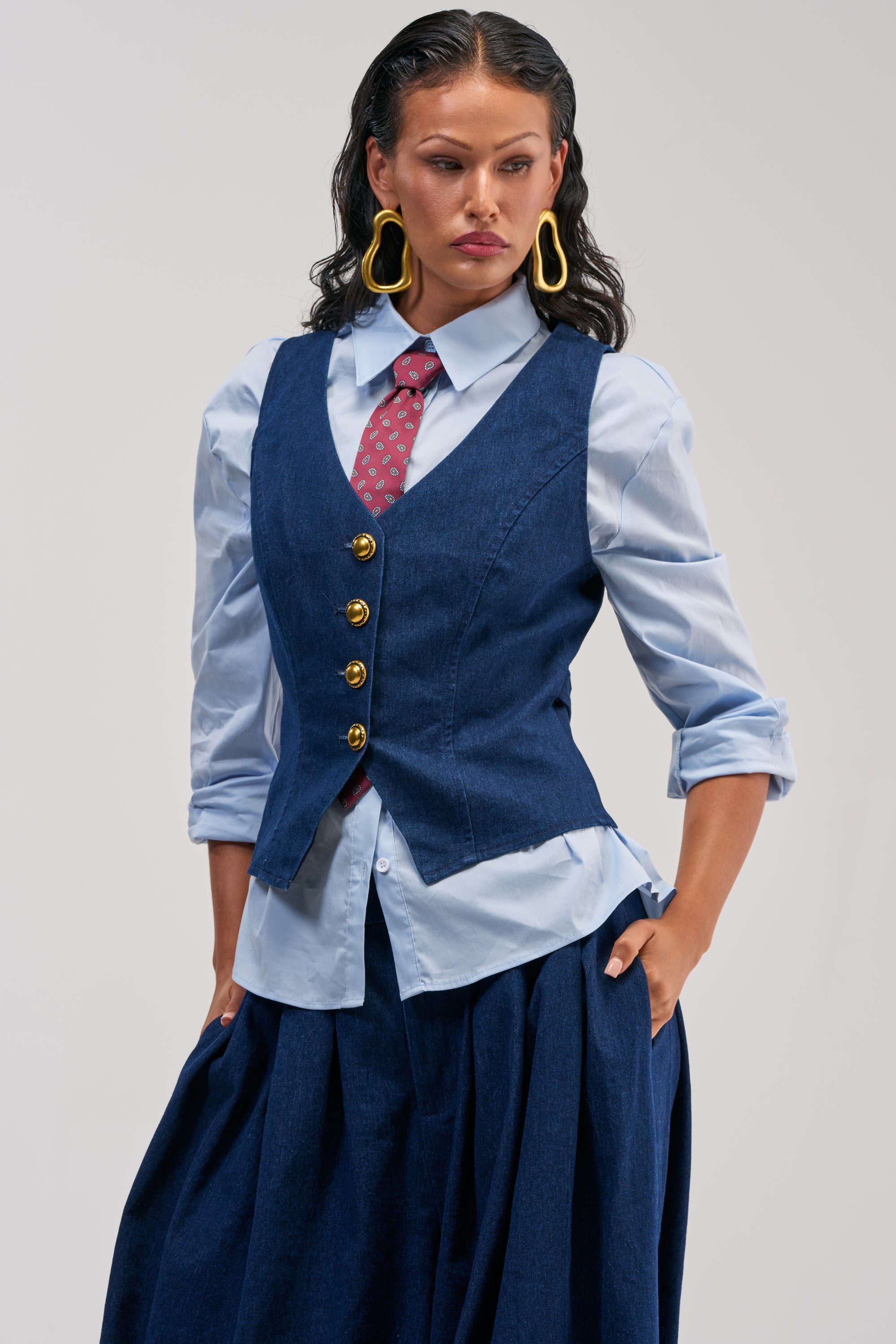 A woman showcases smart-casual style by wearing the BUSINESS IN THE FRONT VEST—a tailored denim piece—layered over a light blue shirt with rolled-up sleeves, finished with a red patterned tie, navy skirt, and bold yellow earrings, as she stands confidently with one hand in her pocket.