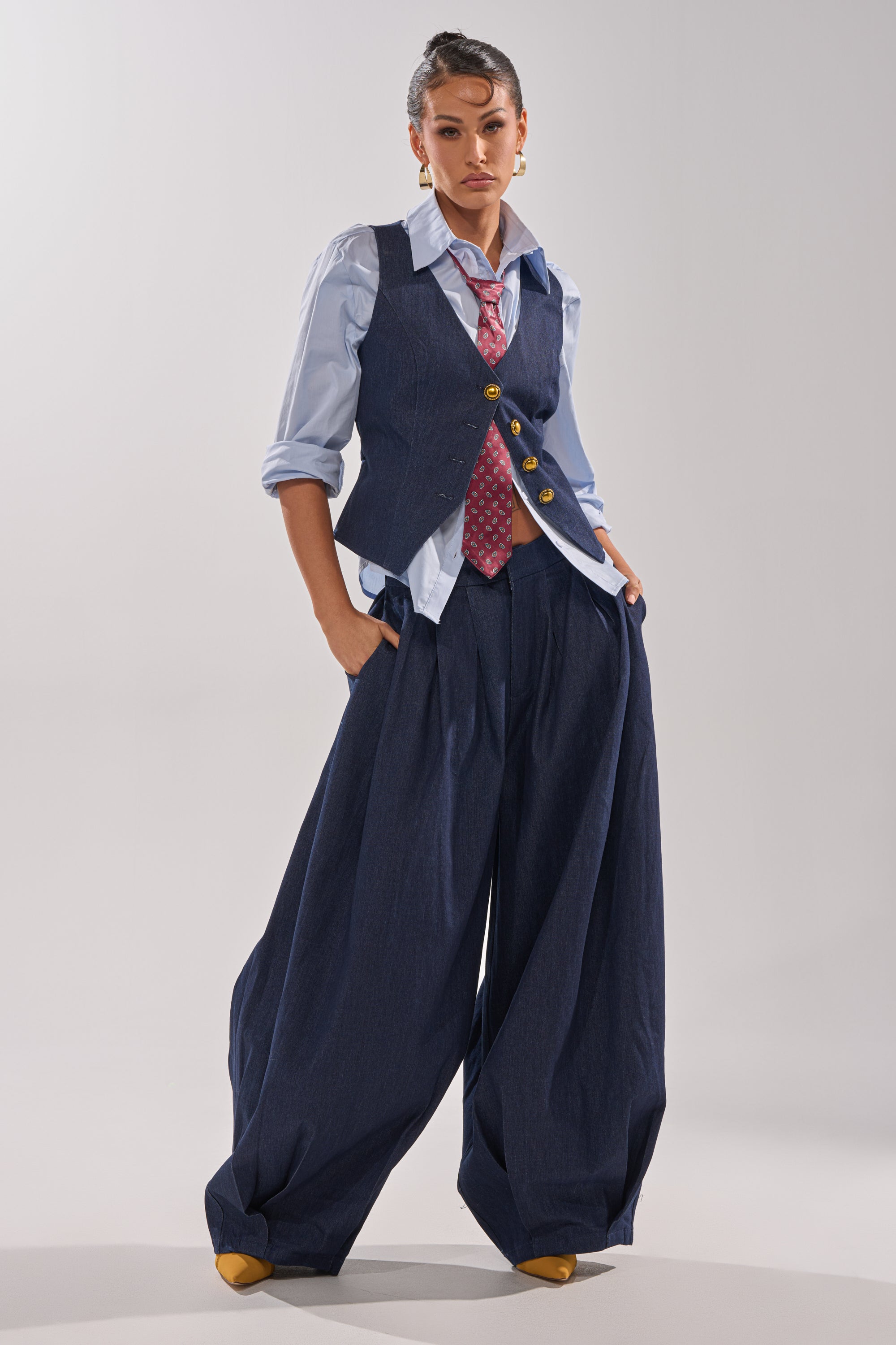 A person stands with hands in pockets, wearing the BUSINESS IN THE FRONT VEST over a blue button-up shirt, paired with oversized wide-leg navy trousers, a red patterned tie, and yellow shoes against a plain light background.