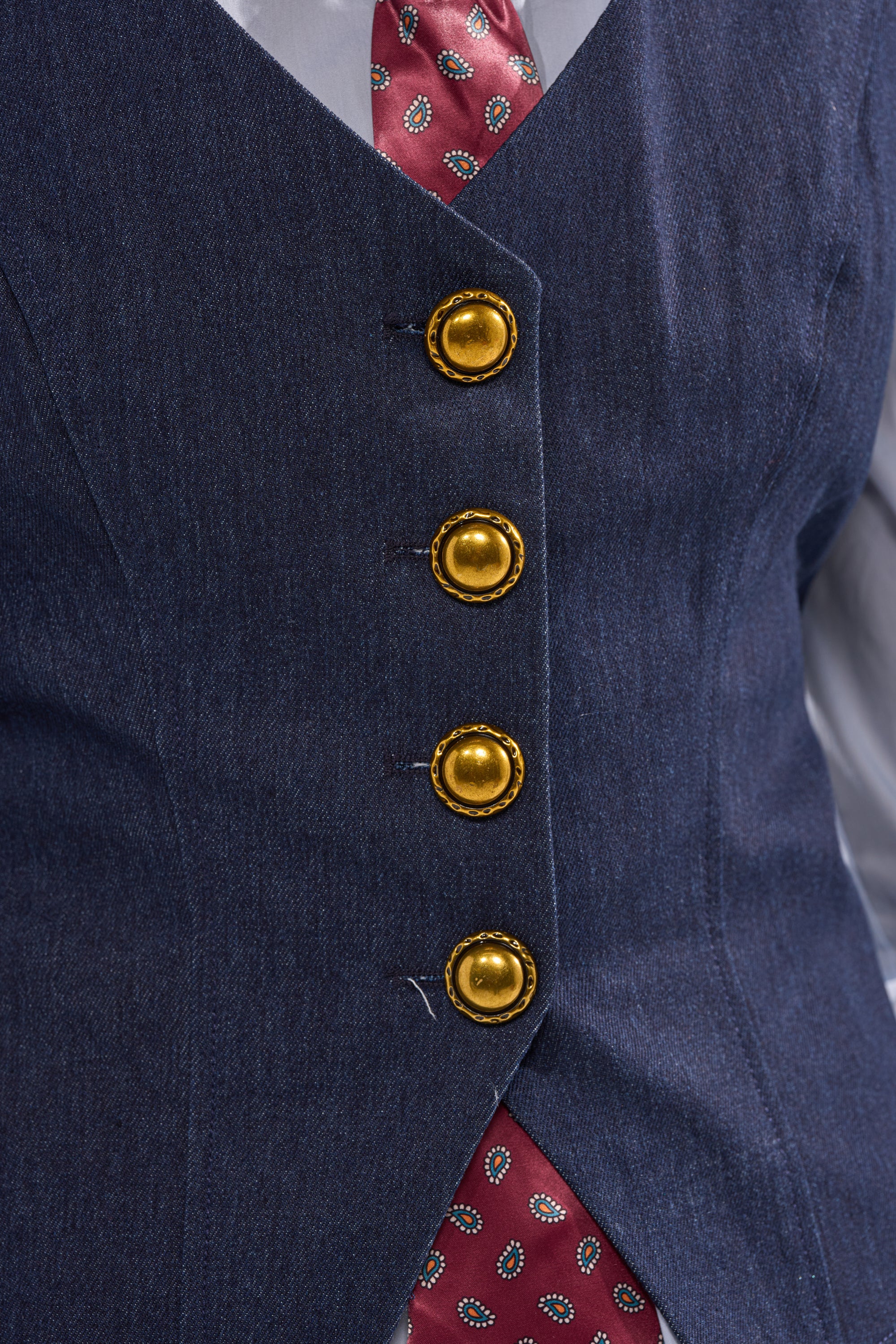 A close-up of a person wearing the BUSINESS IN THE FRONT VEST, a blue tailored piece with four shiny gold buttons, paired with a red patterned tie.