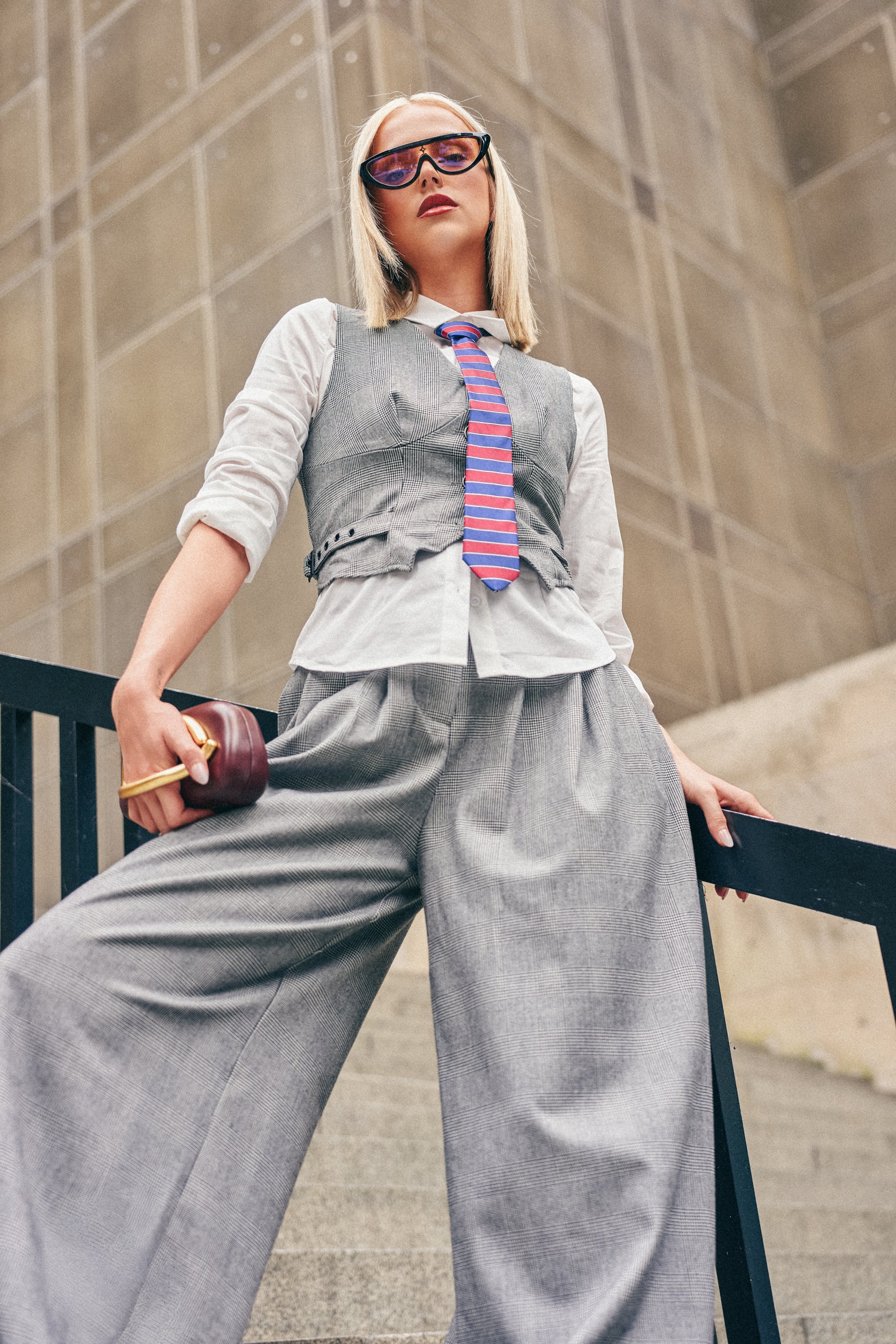 A person in gray wide-leg trousers, the WORK HARD PLAY HARD VEST, a white shirt, and striped tie stands confidently on stairs, holding a clutch and wearing sunglasses—effortlessly showcasing fall streetwear against the backdrop of a modern building.