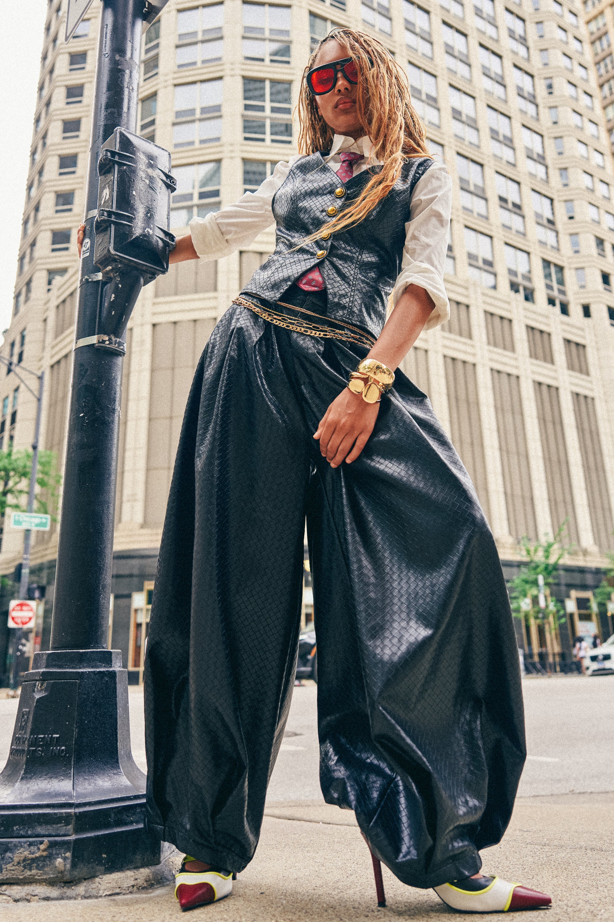 Wearing the OFFICE BADDIE WOVEN PU BALLOON PANT, a person styled in oversized black faux leather pants, a vest, white shirt, and red sunglasses poses confidently on a city street, leaning on a pole with tall buildings in the background.