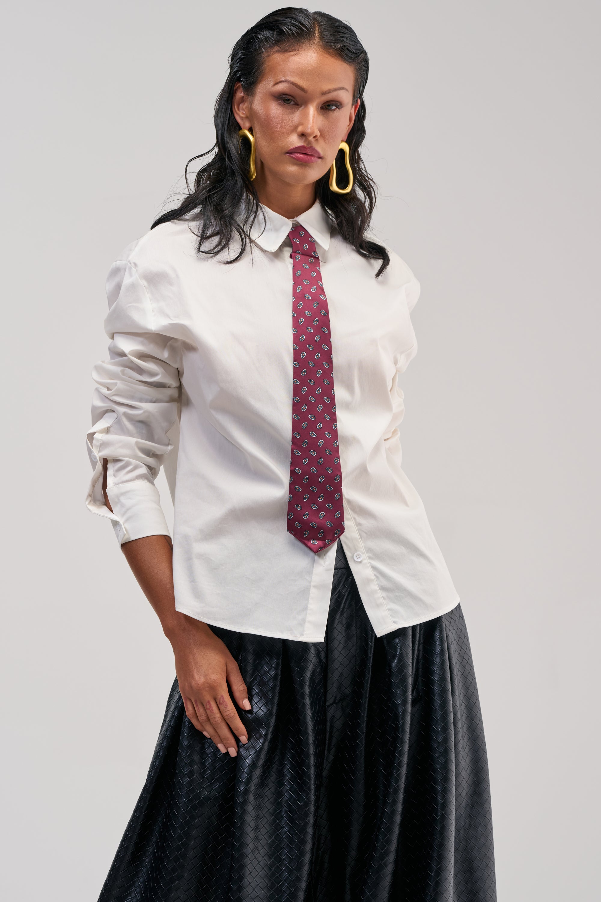 Wearing the OFFICE BADDIE POPLIN BUTTON DOWN BLOUSE with a removable paisley tie, paired with a black skirt and large yellow hoop earrings, a woman stands confidently against a plain background.