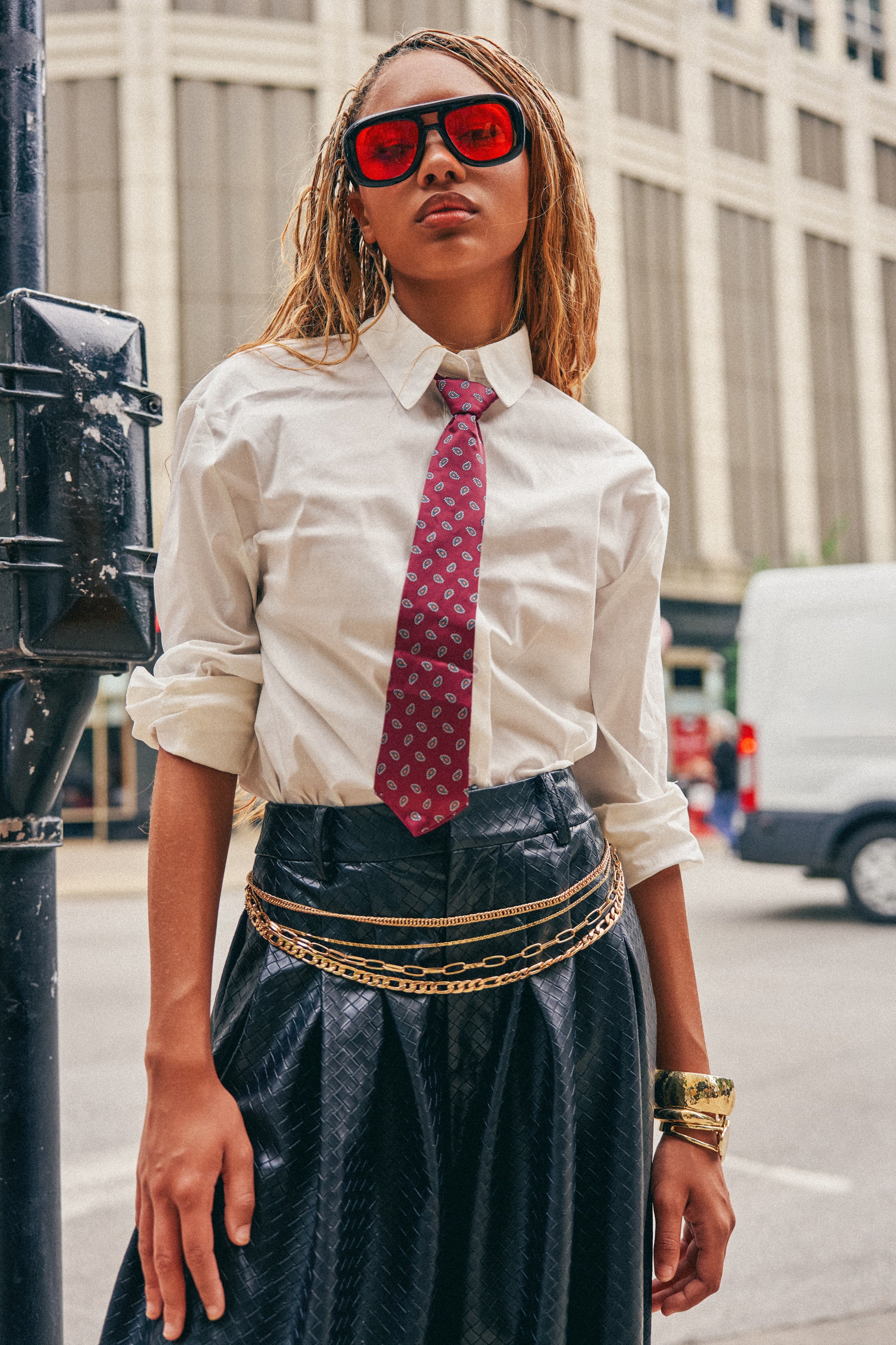 Wearing the OFFICE BADDIE POPLIN BUTTON DOWN BLOUSE, a stylish woman pairs it with a maroon paisley tie, black skirt adorned with gold chains, chunky bracelets, and red sunglasses as she stands confidently on a city street, exuding bold streetwear style.
