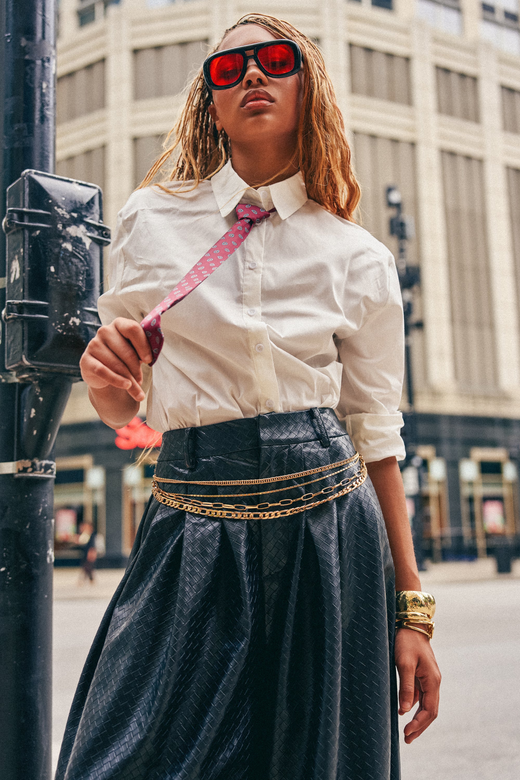 Wearing the OFFICE BADDIE POPLIN BUTTON DOWN BLOUSE, a maroon paisley tie, black skirt with gold chains, sunglasses, and gold bracelets, this person poses confidently on a city street, embodying bold streetwear style with a tall building in the background.