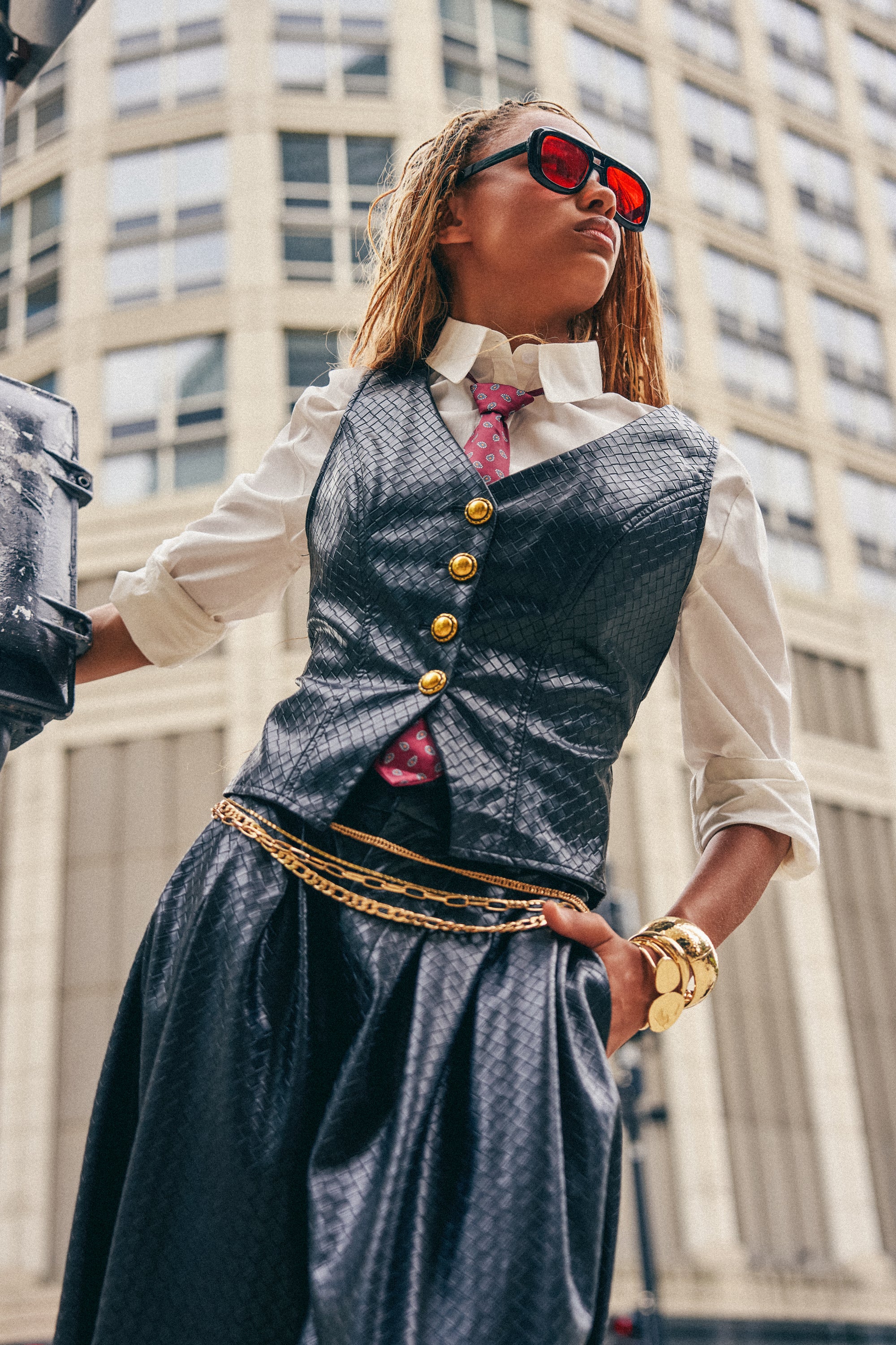 A stylish person in sunglasses, a white shirt, the OFFICE BADDIE WOVEN PU VEST featuring black faux leather with woven construction and gold hardware, a pink polka dot tie, and gold accessories stands confidently on a city street with a tall building in the background.