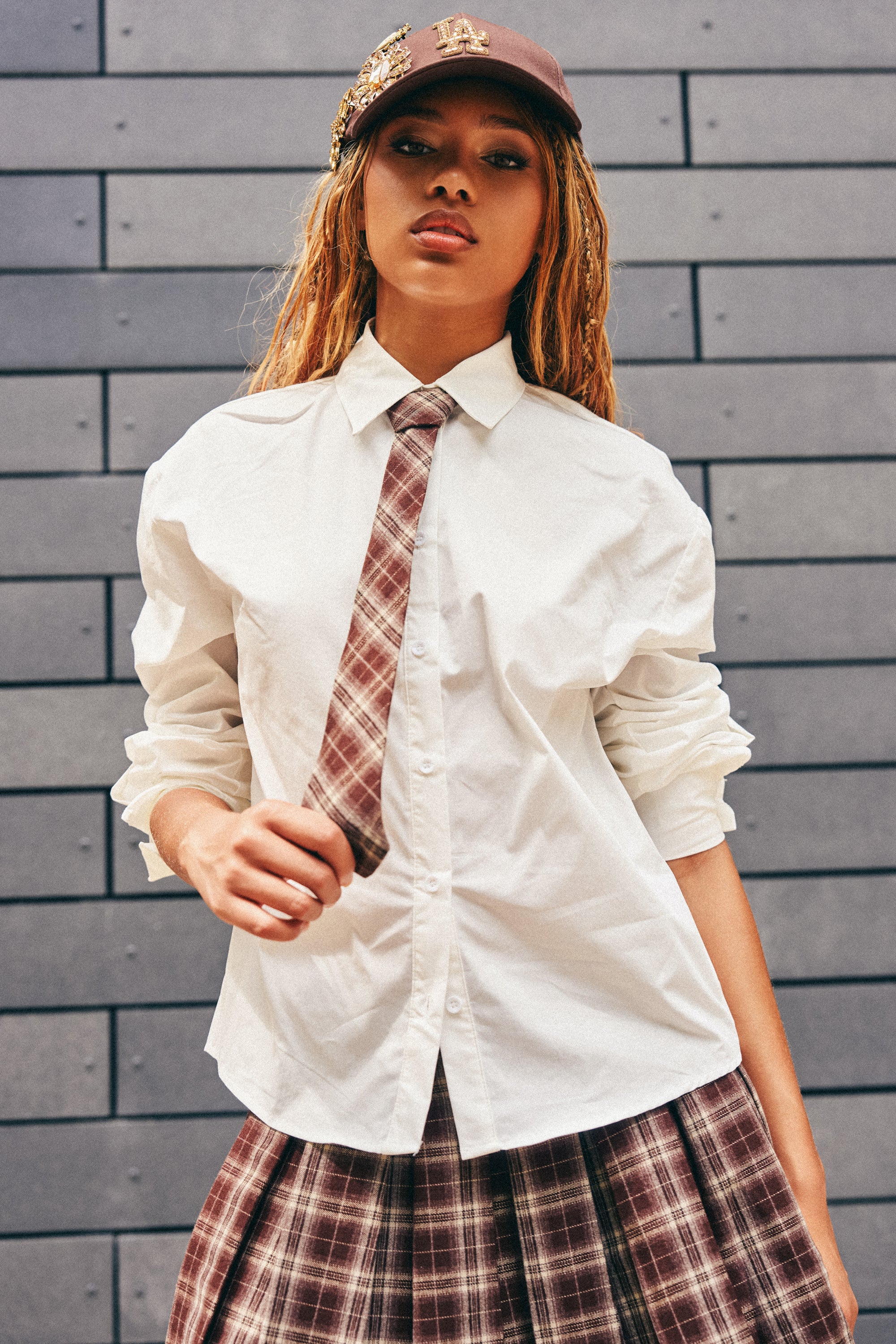Wearing the SCHOOL GIRL VIBES BUTTON DOWN paired with a plaid tie and matching skirt, a person stands in front of a gray tiled wall, topped off with a brown cap.