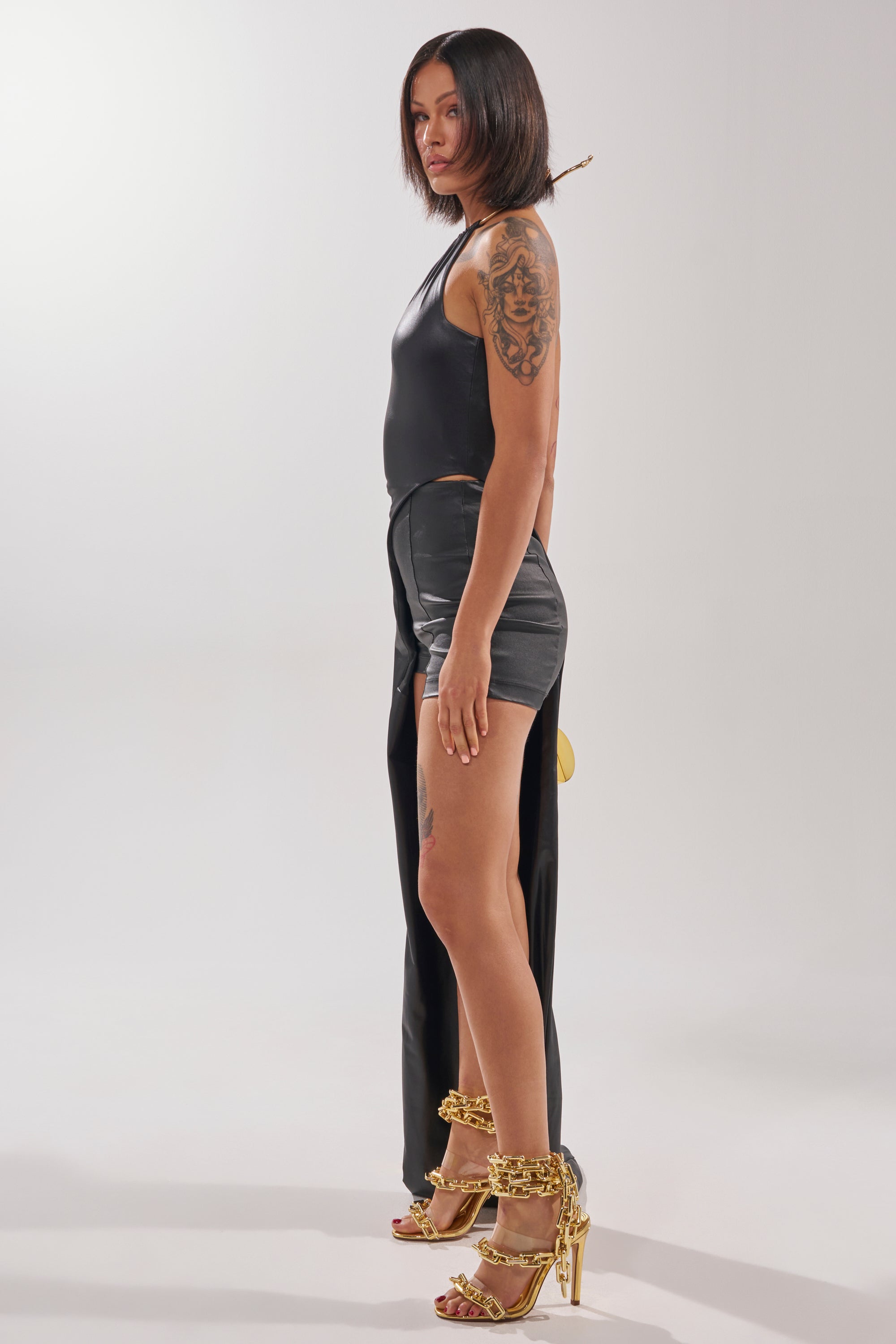 A woman with a bob haircut stands in profile, wearing the DRAMA QUEEN HIGH LOW BLOUSE in black paired with high-waisted shorts and gold heels. She has a visible arm tattoo and a neutral expression.