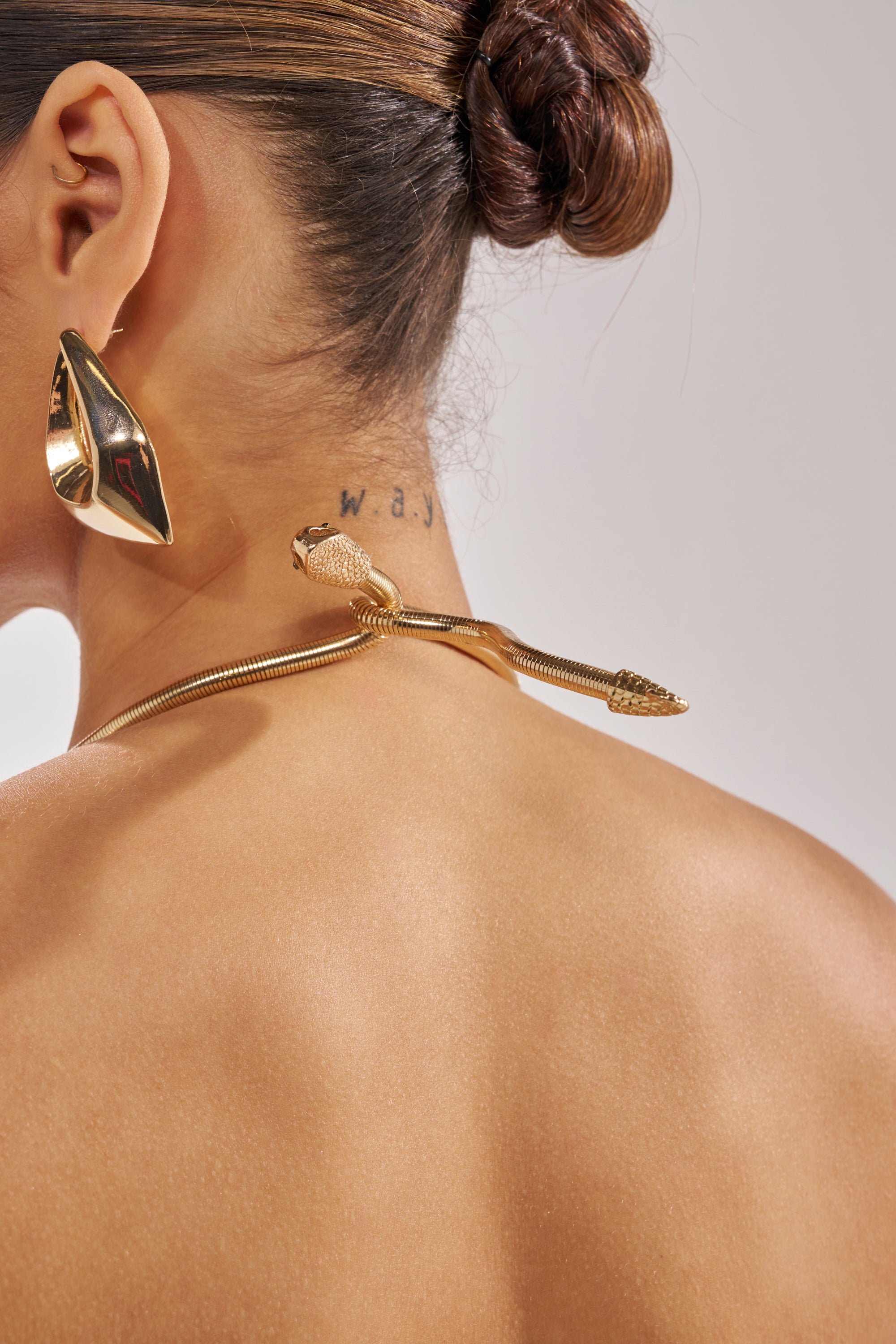A woman with her hair in a bun wears a gold snake necklace, a bold geometric gold earring, and the DRAMA QUEEN HIGH LOW BLOUSE. A "w.a.j" tattoo is visible on her neck against a plain, neutral background. 8