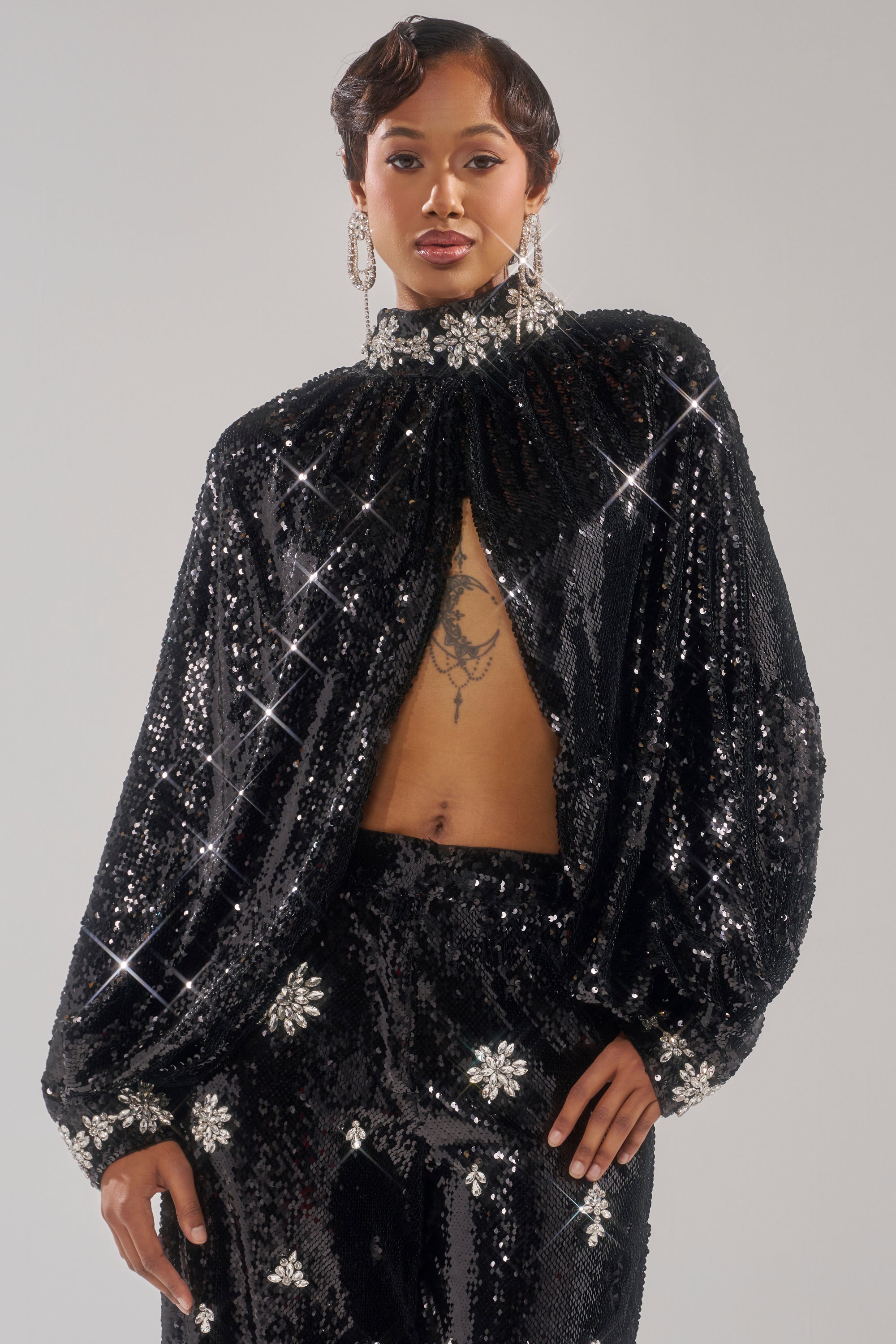 Wearing the DAZZLING HOLIDAY SEQUIN TOP—a black rhinestone cape blouse with wide sleeves, floral sequin accents, and a high neck—they reveal part of their chest, a tattoo, short styled hair, and bold earrings.