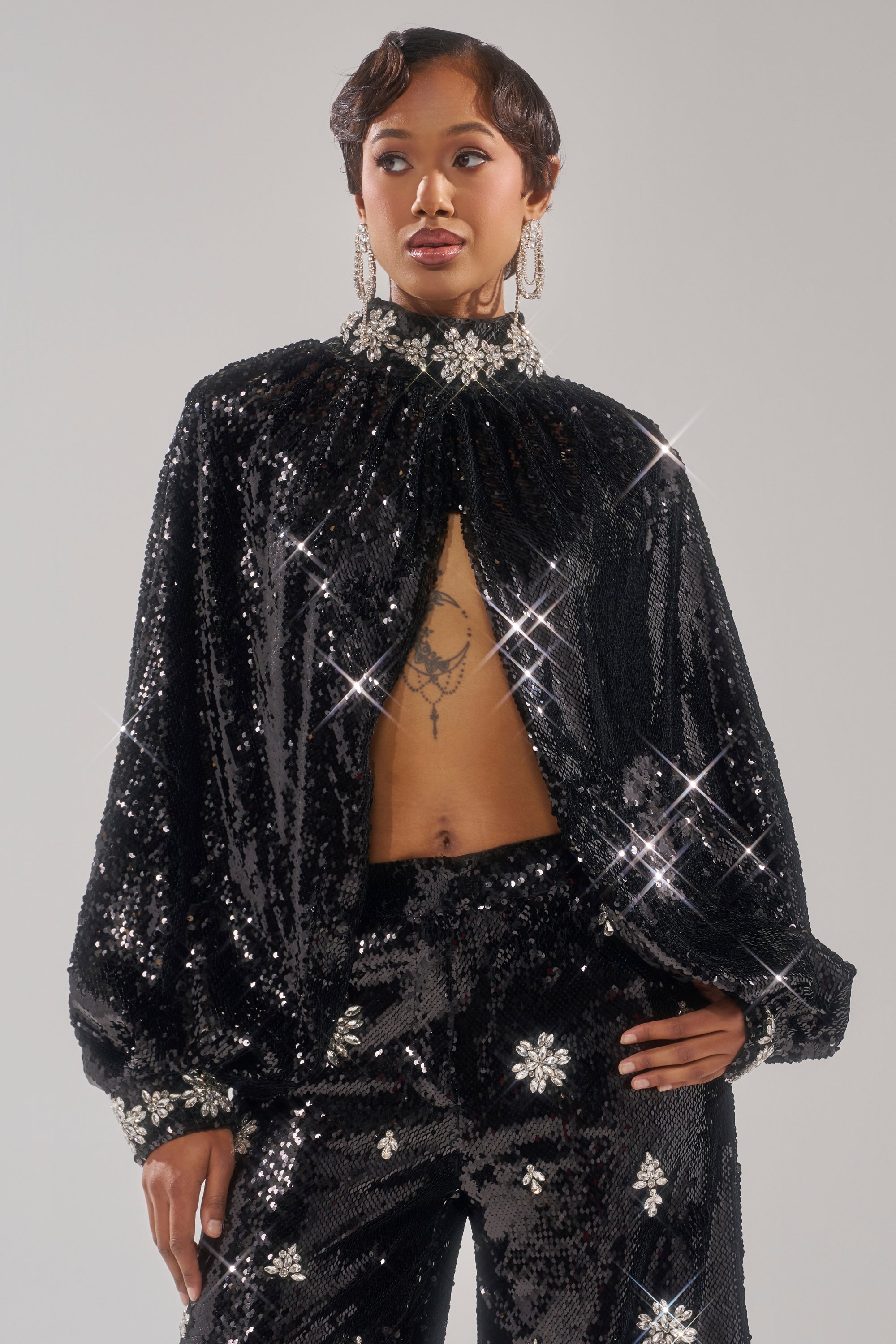 Wearing the DAZZLING HOLIDAY SEQUIN TOP—sparkling black with silver floral accents, wide sleeves, and an open front—a person stands confidently against a plain gray background.
