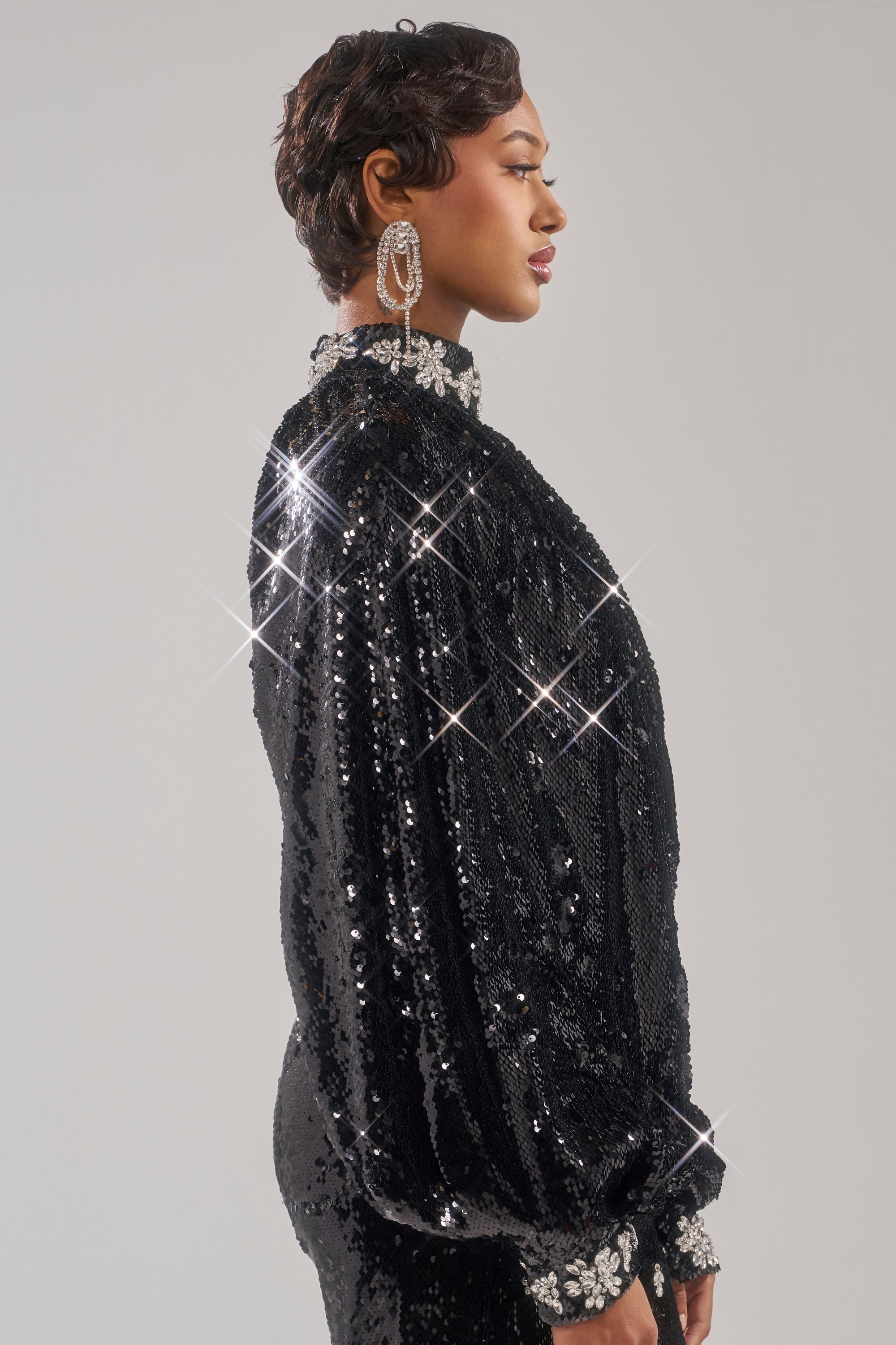 A person with short hair, in profile against a plain light background, wears large sparkling earrings and the DAZZLING HOLIDAY SEQUIN TOP with jeweled collar and cuffs. Shimmering sequins make this festive Christmas party outfit stand out.