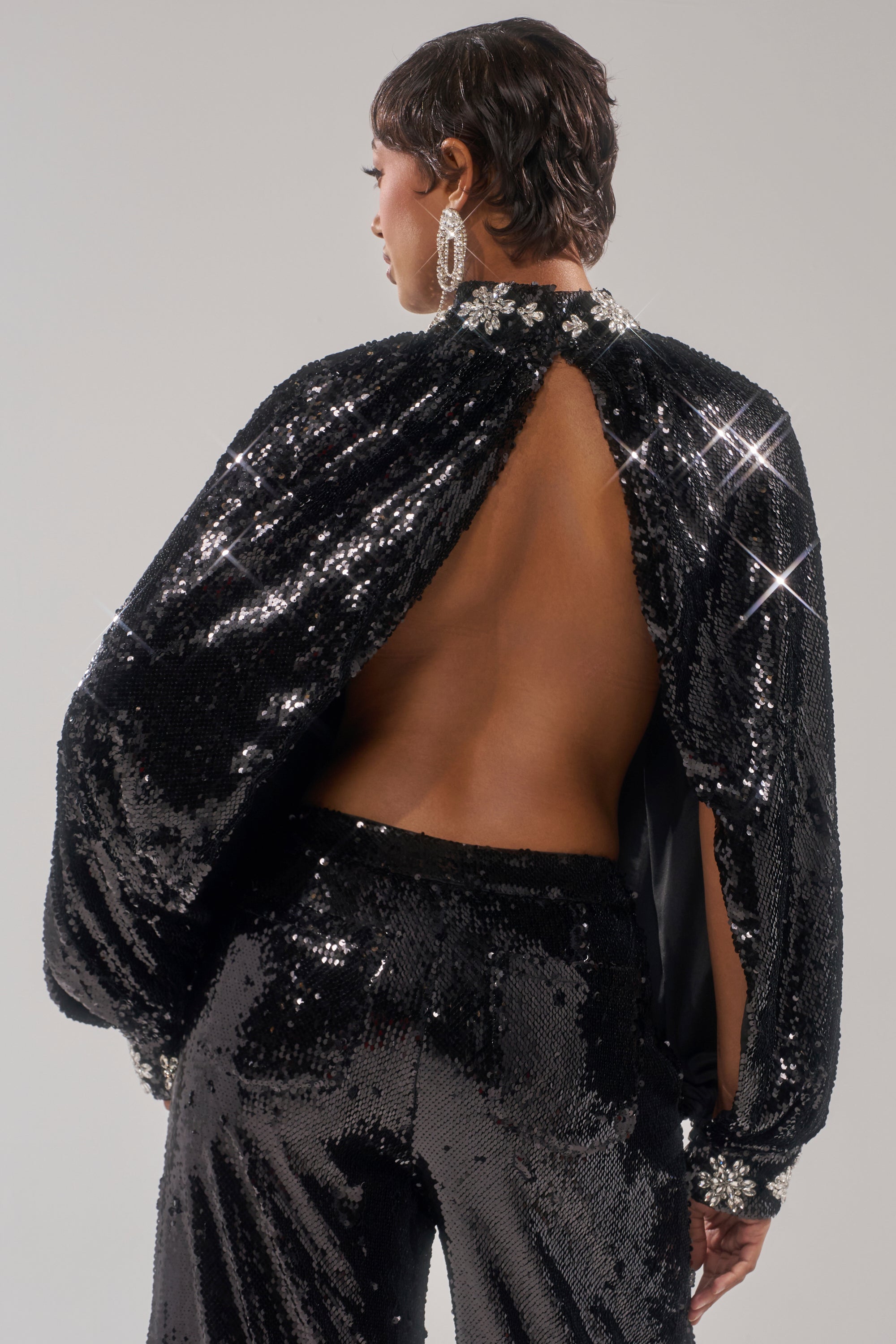 A person with short hair wears large hoop earrings and the DAZZLING HOLIDAY SEQUIN TOP, featuring an open back and sparkling black rhinestones, standing against a plain background—perfect for a festive look.