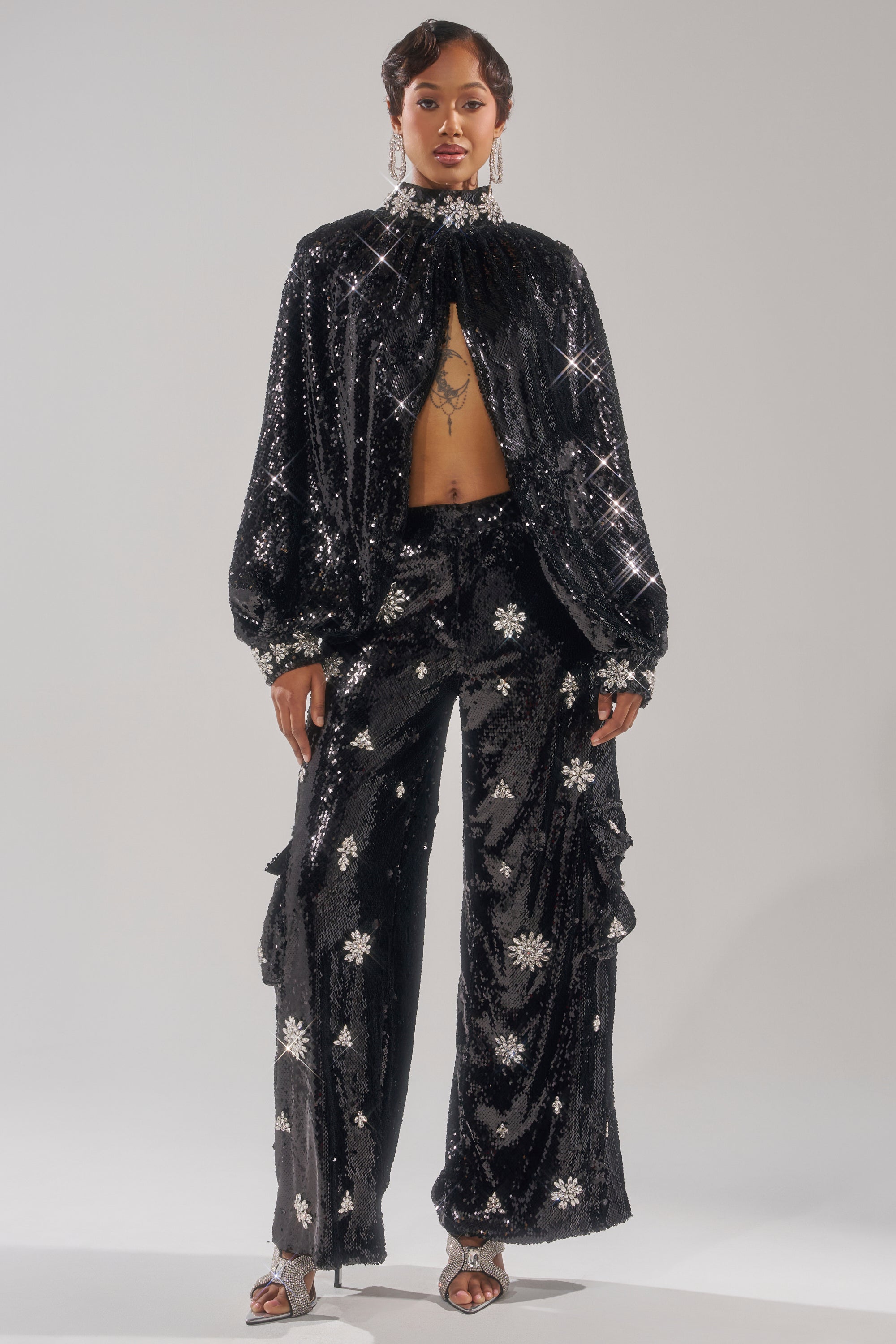 A woman poses confidently in the DAZZLING HOLIDAY SEQUIN TOP and matching wide-leg pants, both black with silver rhinestone starburst patterns. She completes the look with short hair, statement earrings, and silver heels against a plain backdrop.