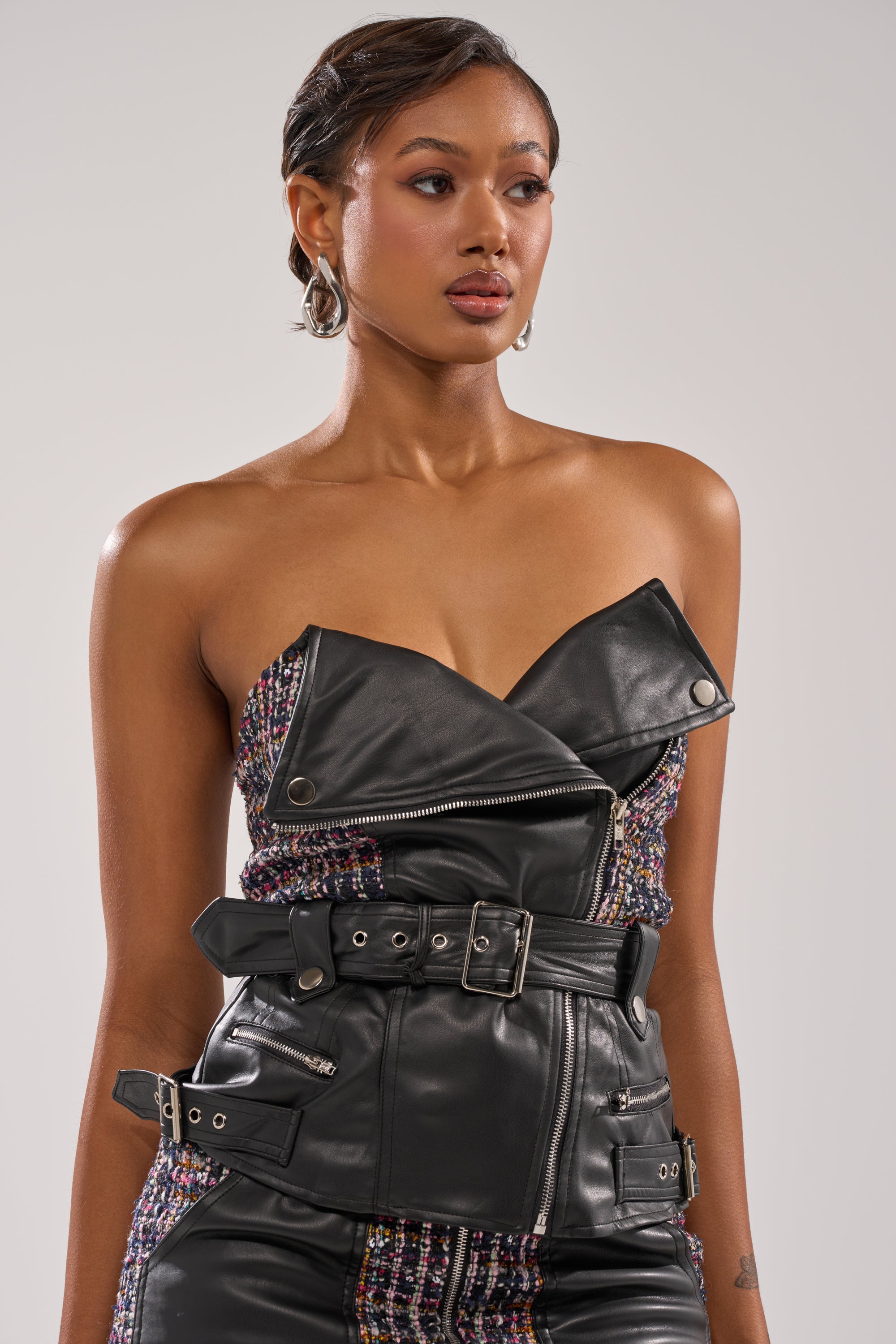 A woman with short hair wears the WHAT YOU GOT FOR ME TWEED CORSET, a strapless crop top with zipper and buckle details and metallic-textured panels. She accessorizes with hoop earrings, looking off to the side against a plain background.