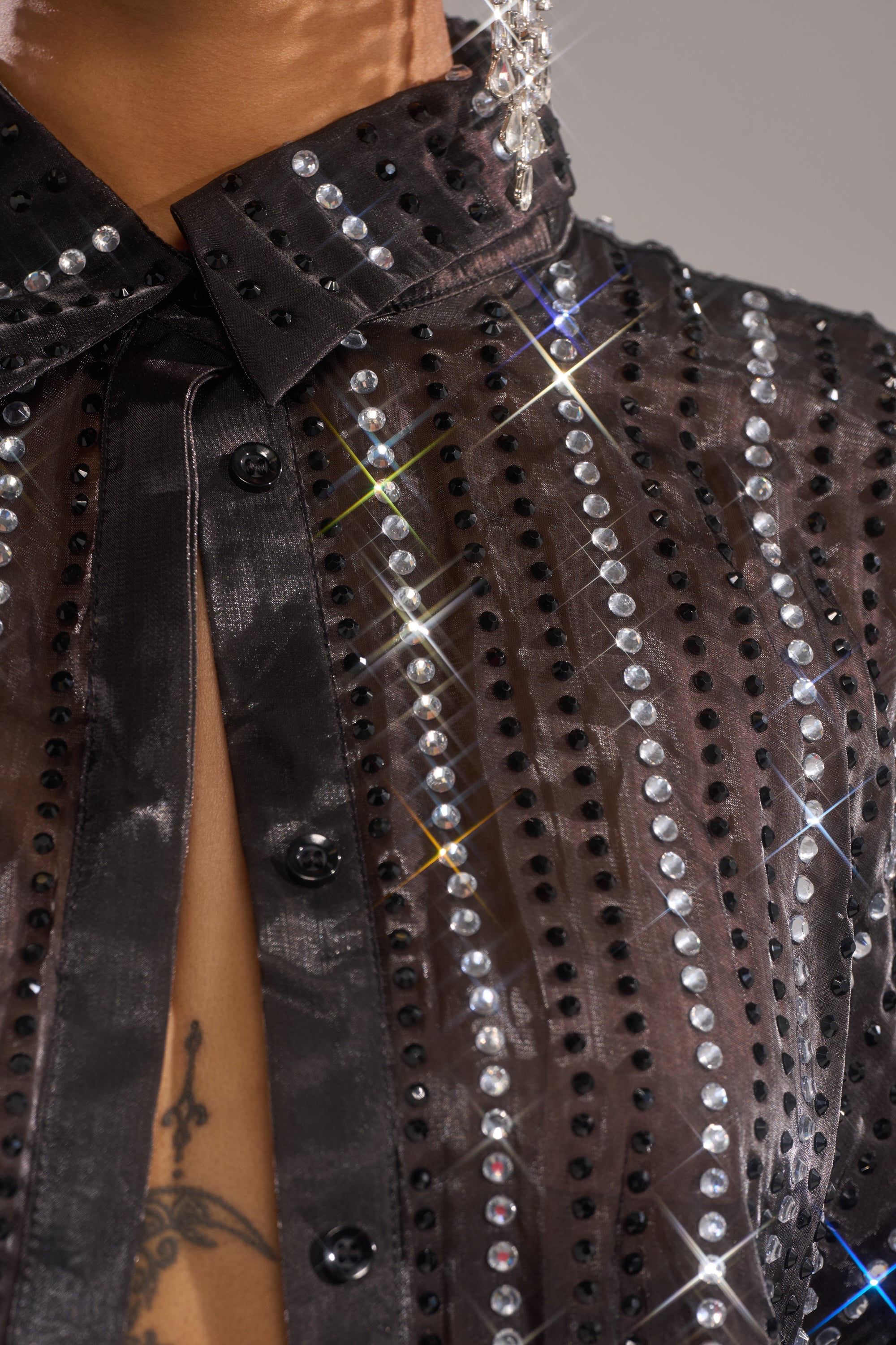 Close-up of the DANCE THE NIGHT AWAY BUTTON DOWN sheer chiffon top with vertical rows of shiny rhinestones and small black studs, reflecting light for a sparkling effect. Visible buttons and part of a tattoo under the shirt.