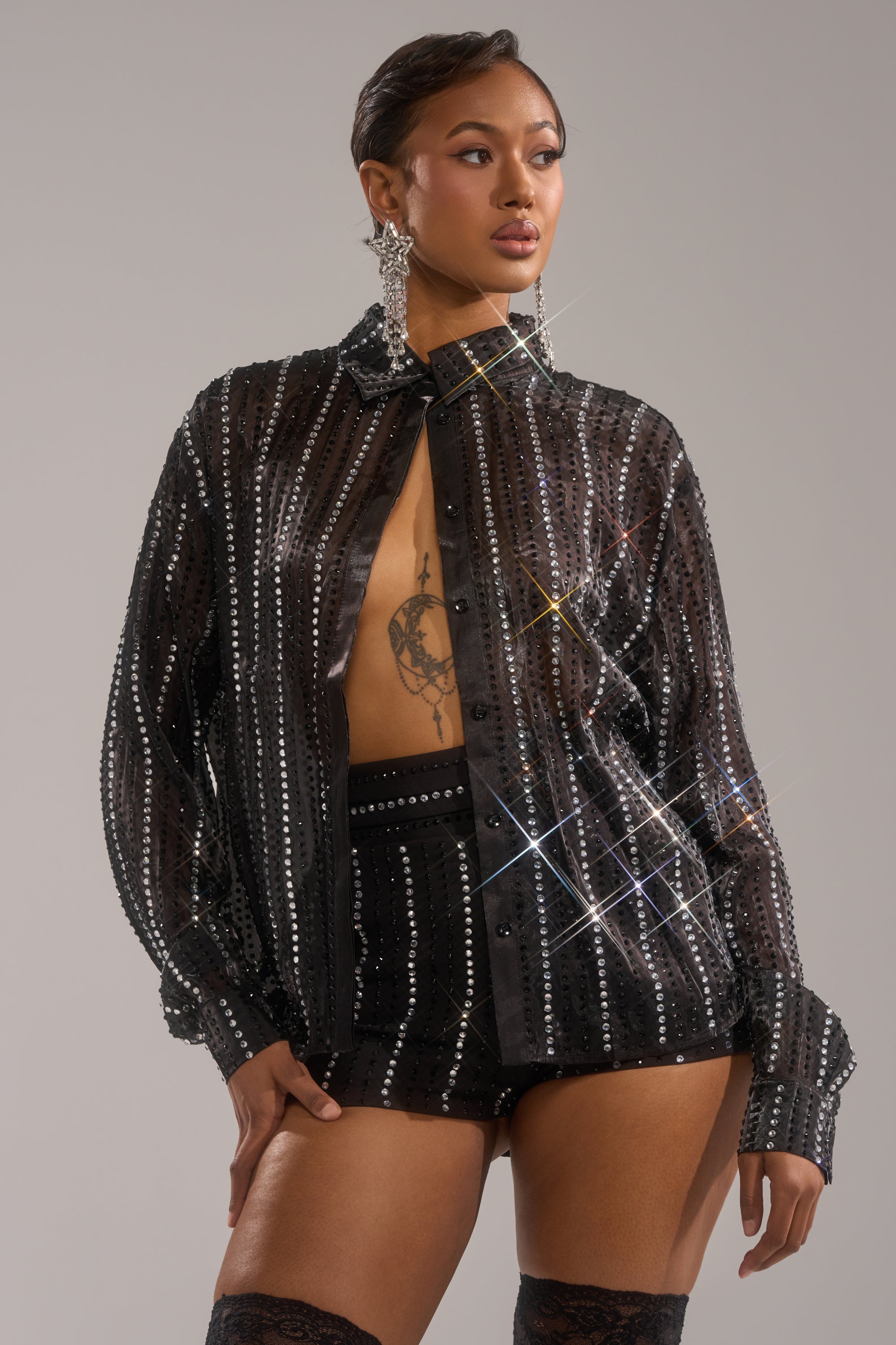 A woman showcases the DANCE THE NIGHT AWAY BUTTON DOWN, a sheer chiffon top with vertical sequins, paired with high-waisted black shorts, sparkly earrings, and thigh-high stockings against a neutral background.