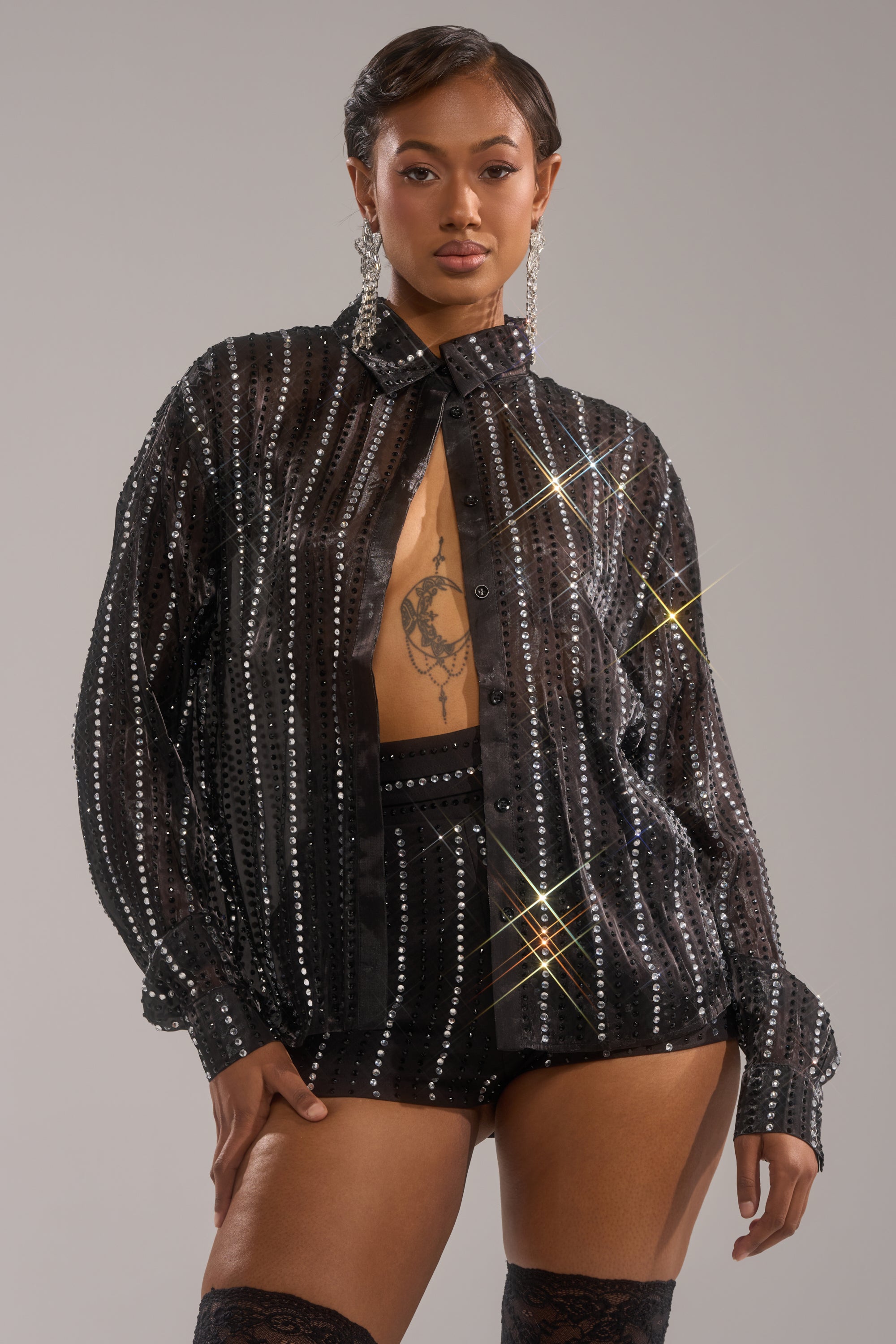 A woman poses confidently in the DANCE THE NIGHT AWAY BUTTON DOWN, a sheer chiffon top with sparkling rhinestone stripes, layered over a black bra and shorts. She sports short hair, statement earrings, and displays a chest tattoo.
