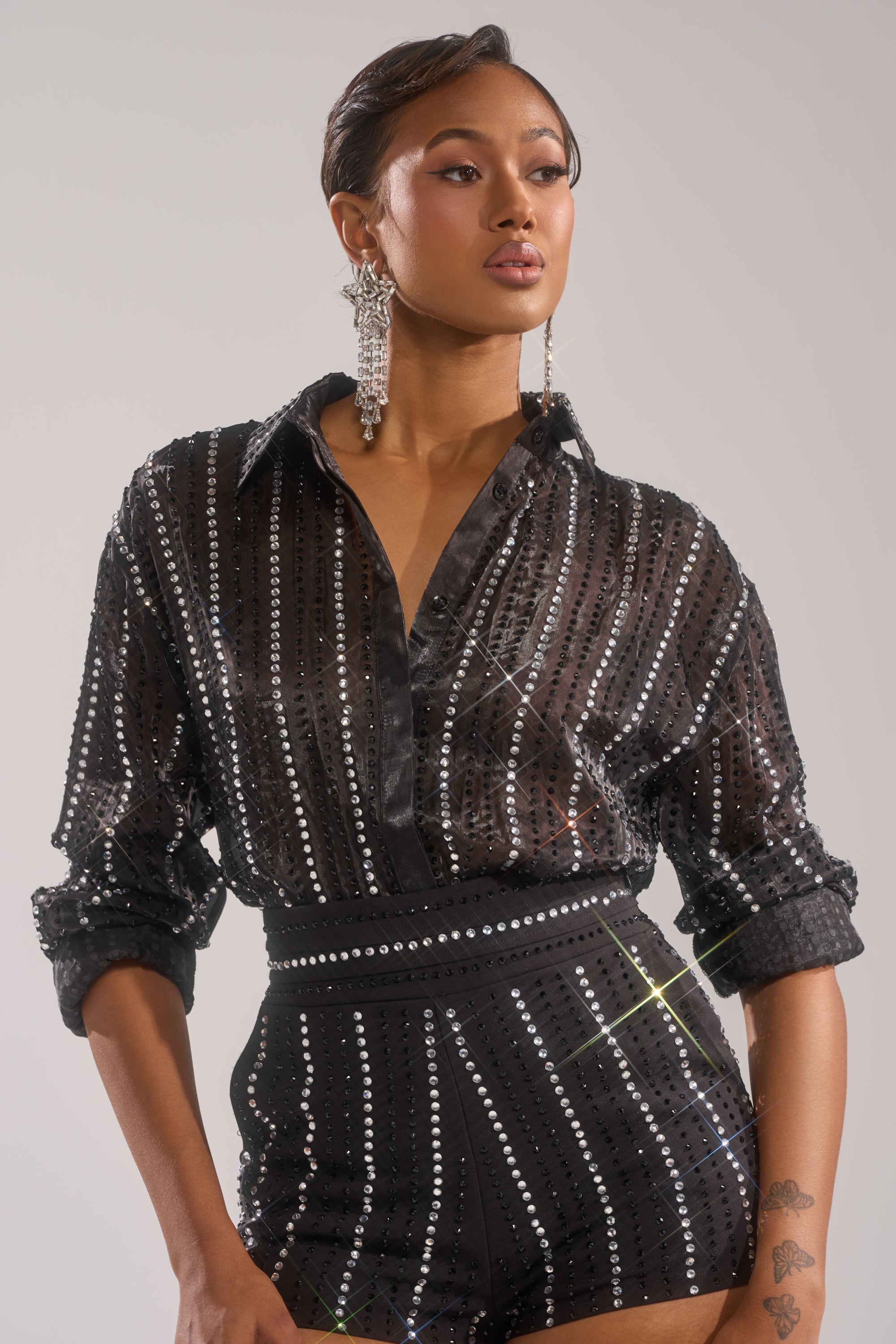 A woman poses in the AKIRA Label DANCE THE NIGHT AWAY BUTTON DOWN sheer chiffon top with matching high-waisted sequin shorts, paired with large dangling earrings and showcasing a floral tattoo on her left forearm.